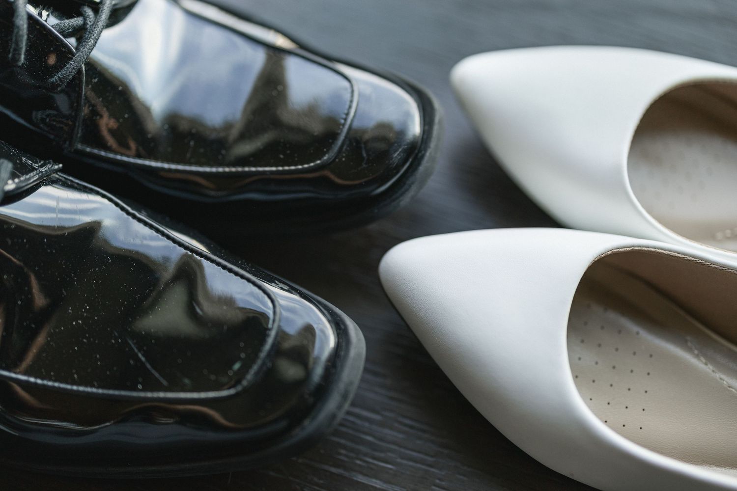 Black leather dress shoes and white pointed heels arranged on dark wooden surface.