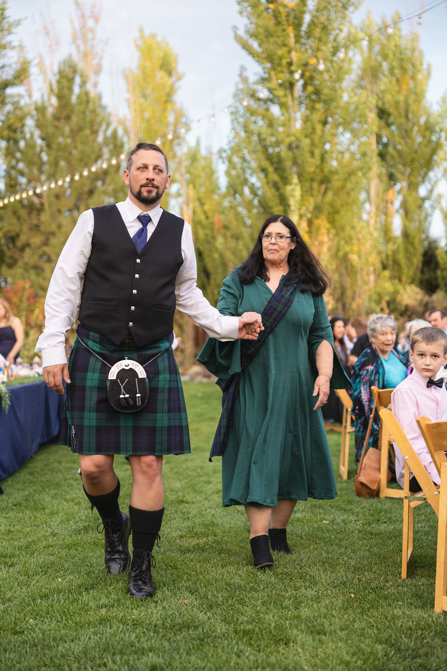 A man in traditional Scottish kilt walks with a woman in green dress at an outdoor wedding ceremony.