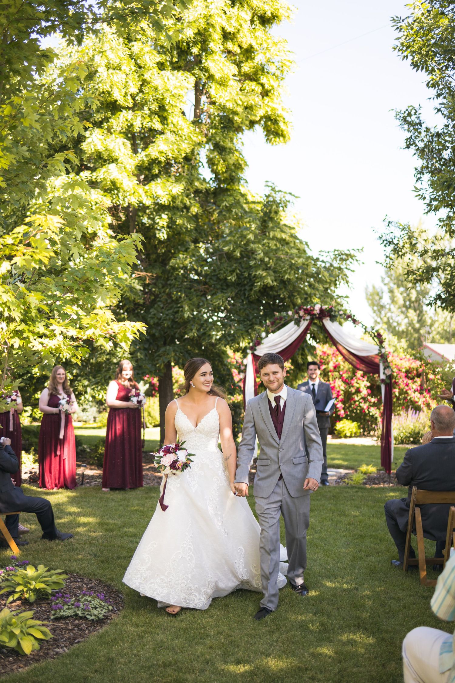 Newlyweds walking down the aisle after ceremony under tall trees and burgundy arch decor.