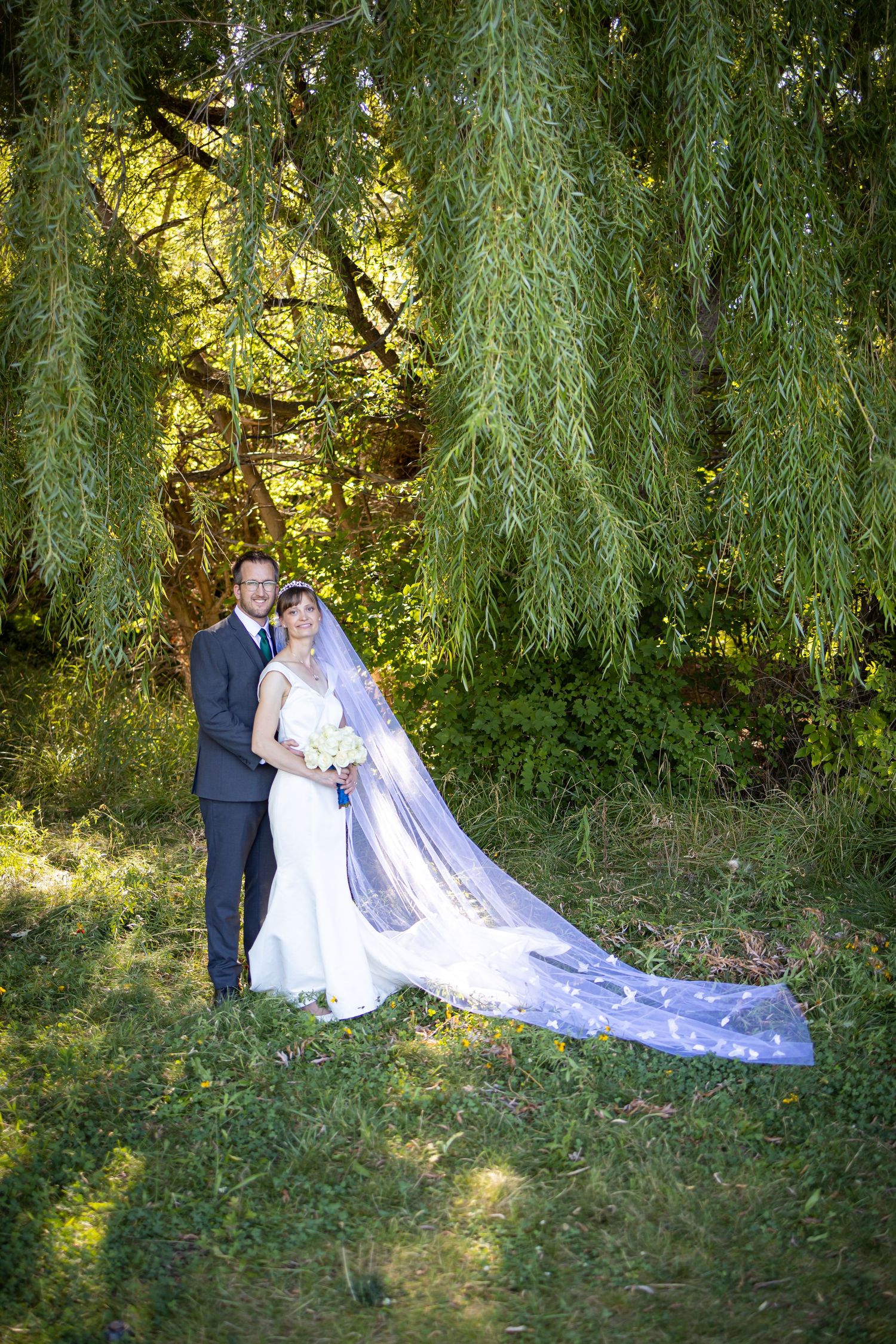 Wedding couple poses under drooping willow tree branches in garden setting.