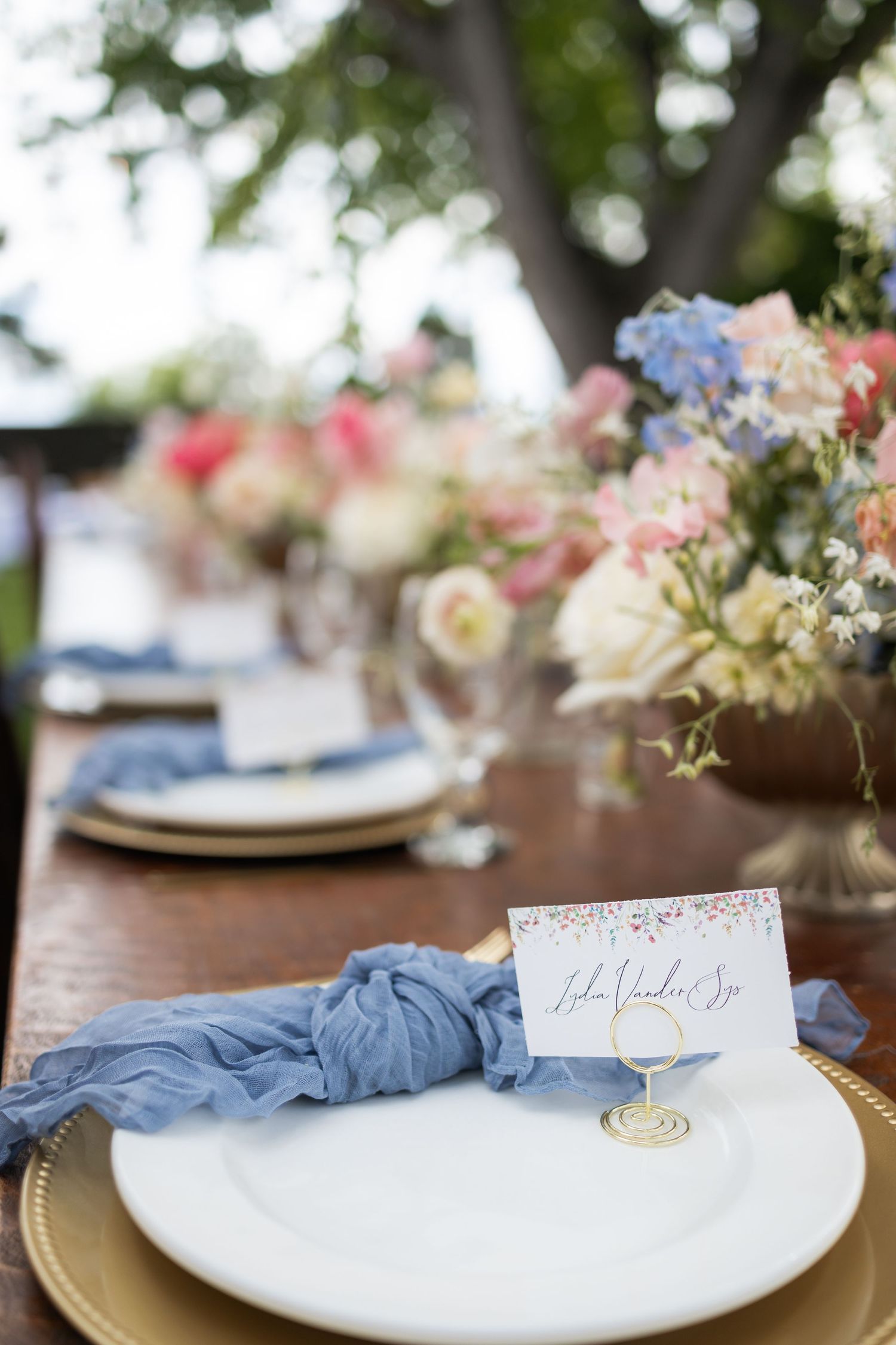 Elegant place setting with blue napkin and name card on gold-rimmed plate with floral centerpiece.
