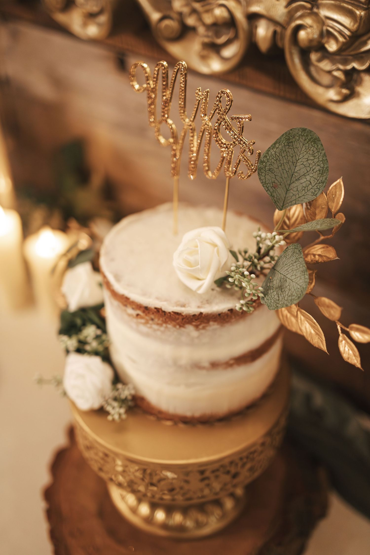 Small white naked wedding cake with gold accents and eucalyptus leaves on vintage gold stand.