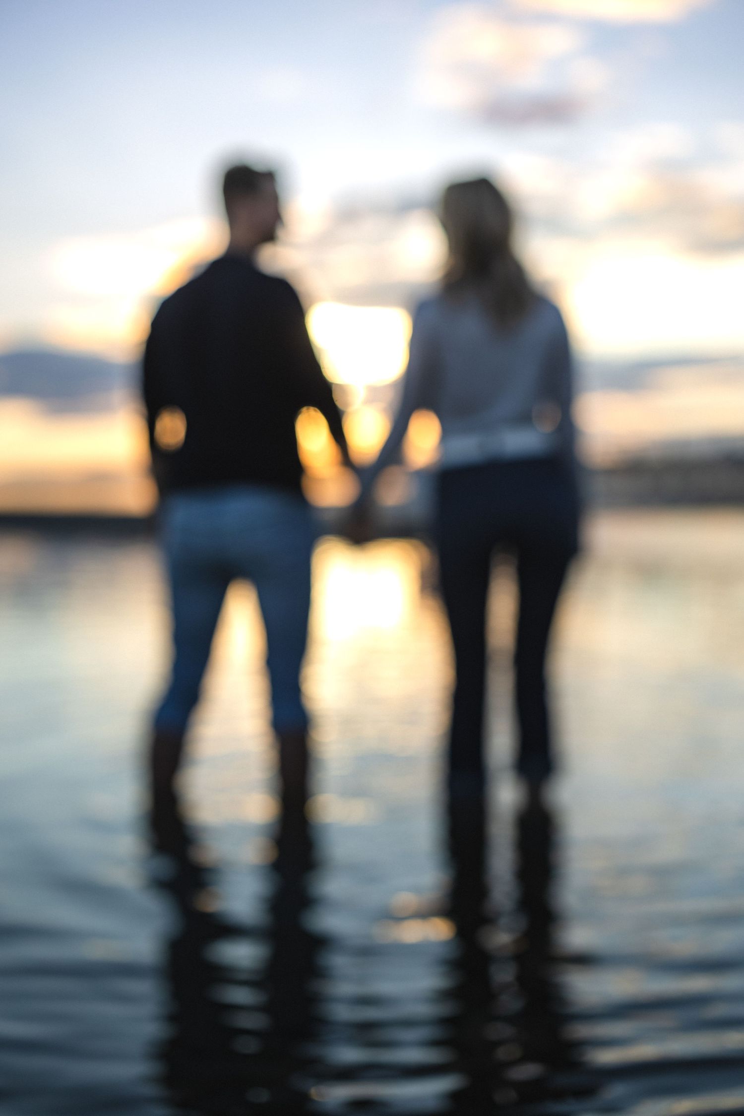Silhouetted couple holding hands while standing in shallow water at sunset.