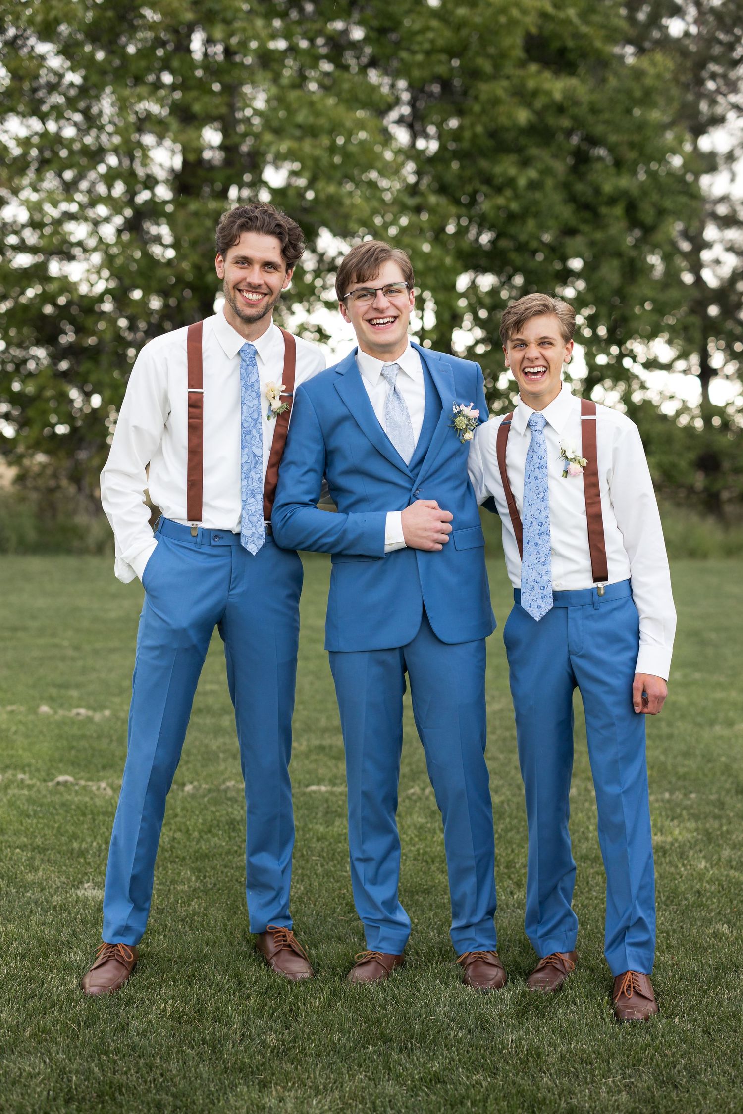 Three groomsmen in coordinating blue suits and brown leather suspenders pose on grass for wedding photos.