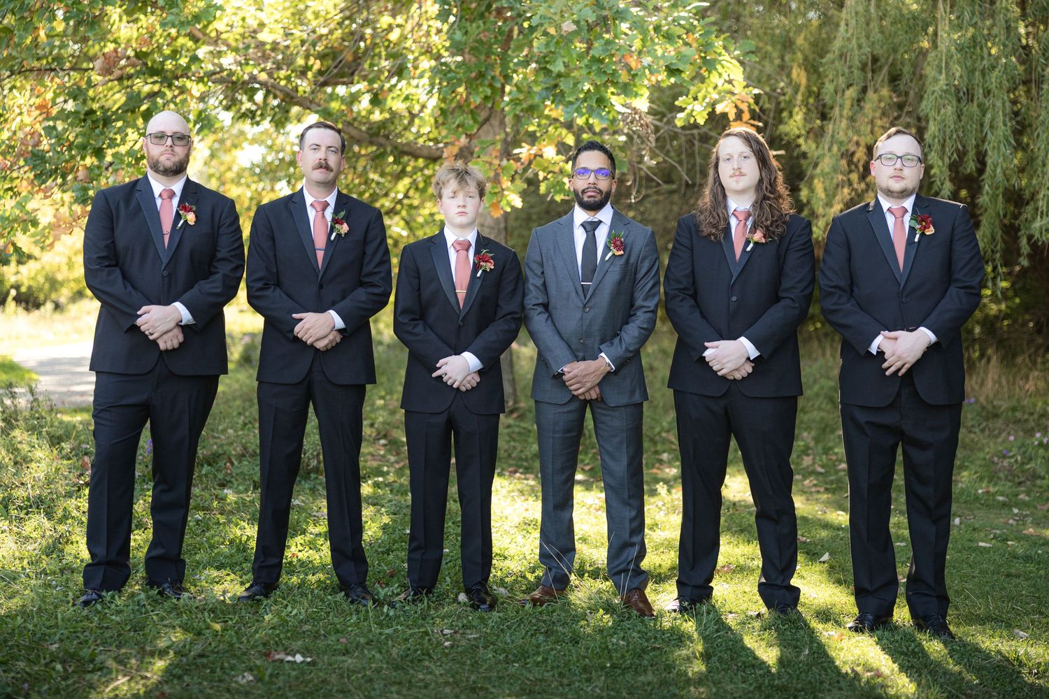 Wedding party dressed in black suits and dusty rose dresses poses together in an outdoor garden setting.