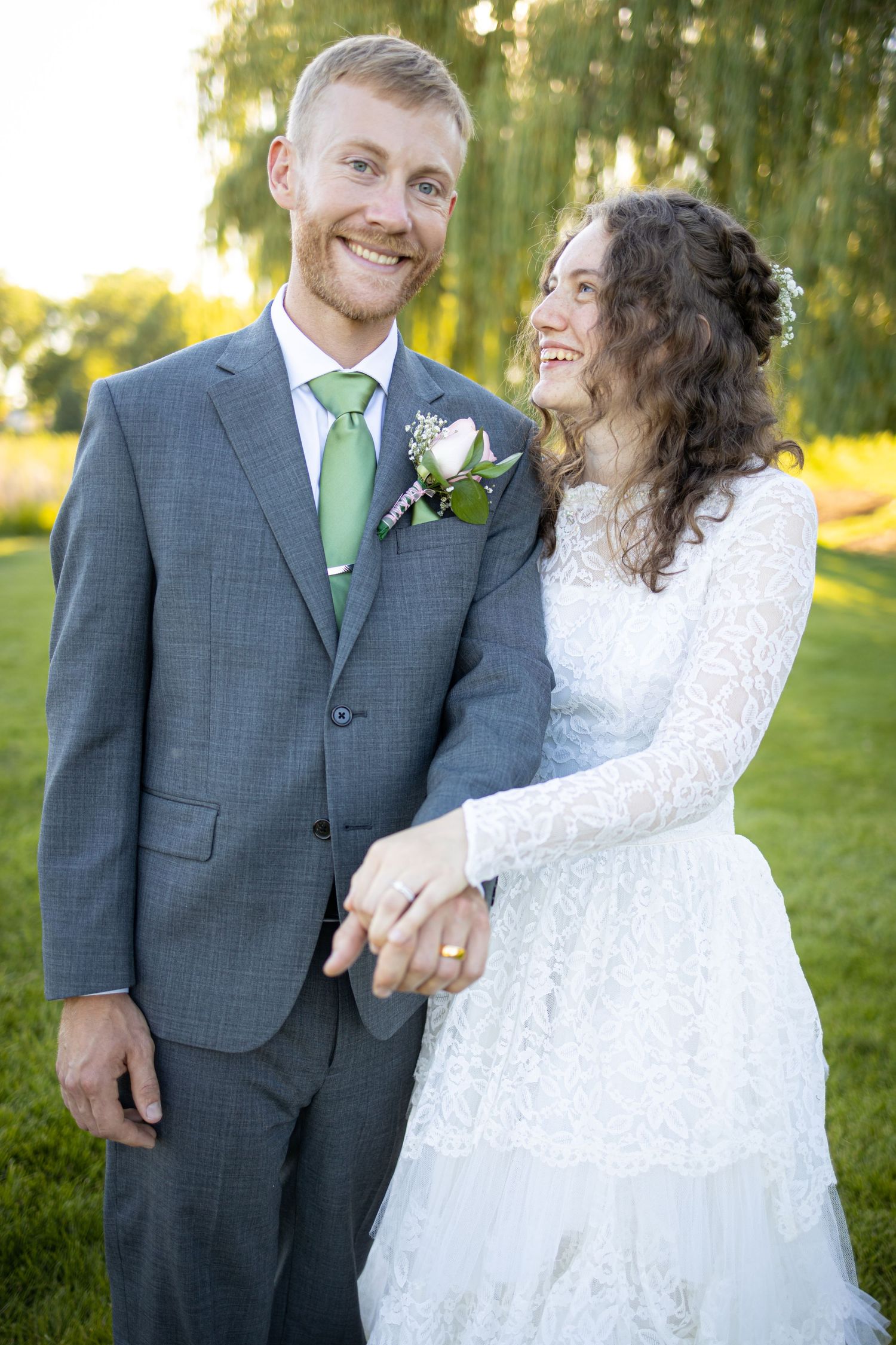 Newlyweds in grey suit and white lace dress smiling together in garden setting with willow trees.