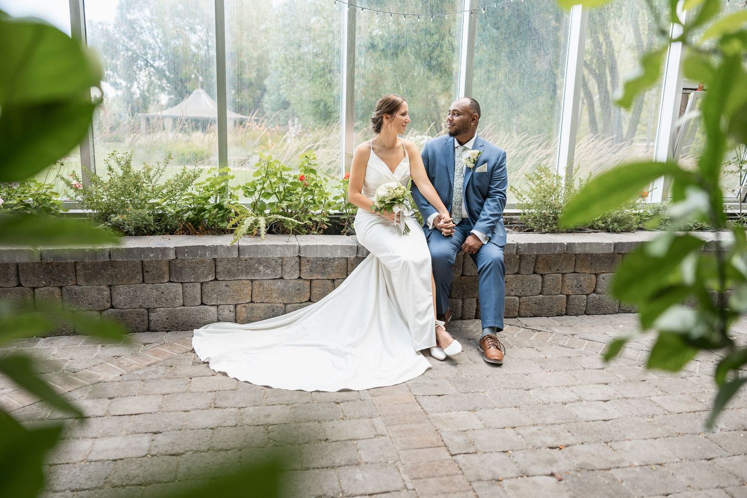 Wedding couple seated on stone wall with greenhouse and garden scenery in background.