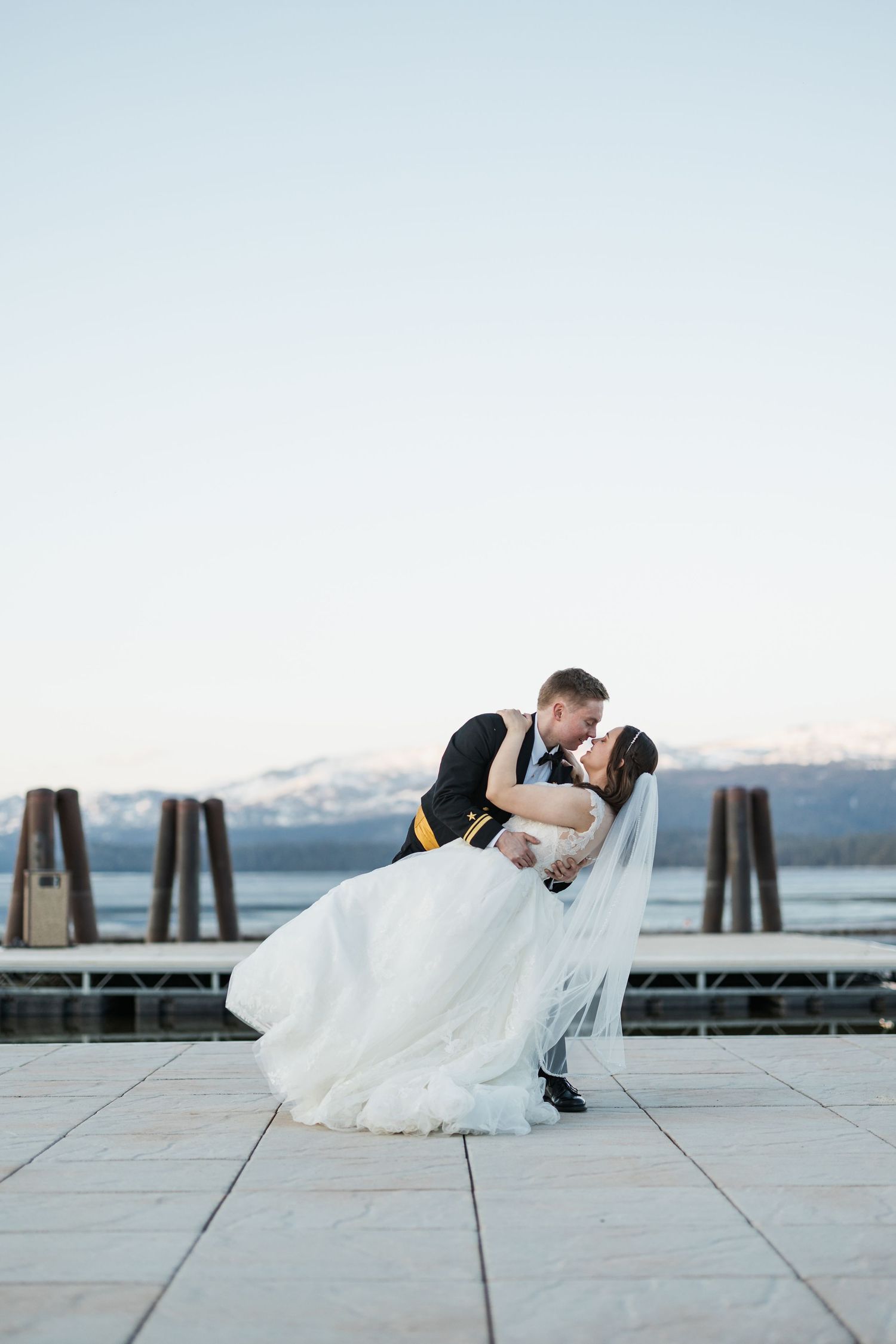 A romantic wedding photo shows a couple sharing a kiss on a dock with mountains and water in the background.