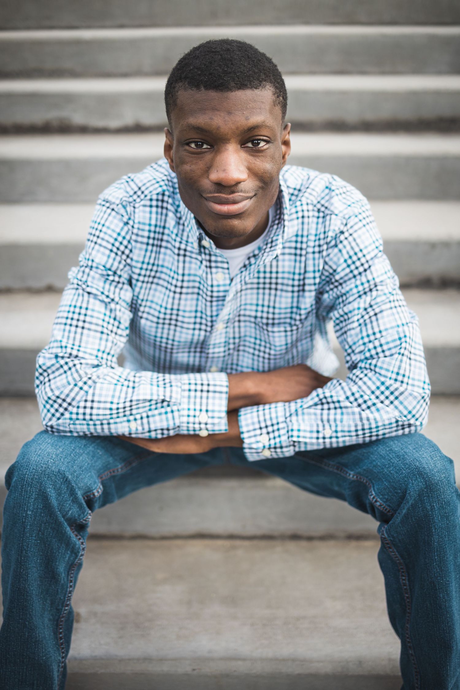 A person in a blue checkered shirt and jeans sits casually on concrete steps with a warm smile.