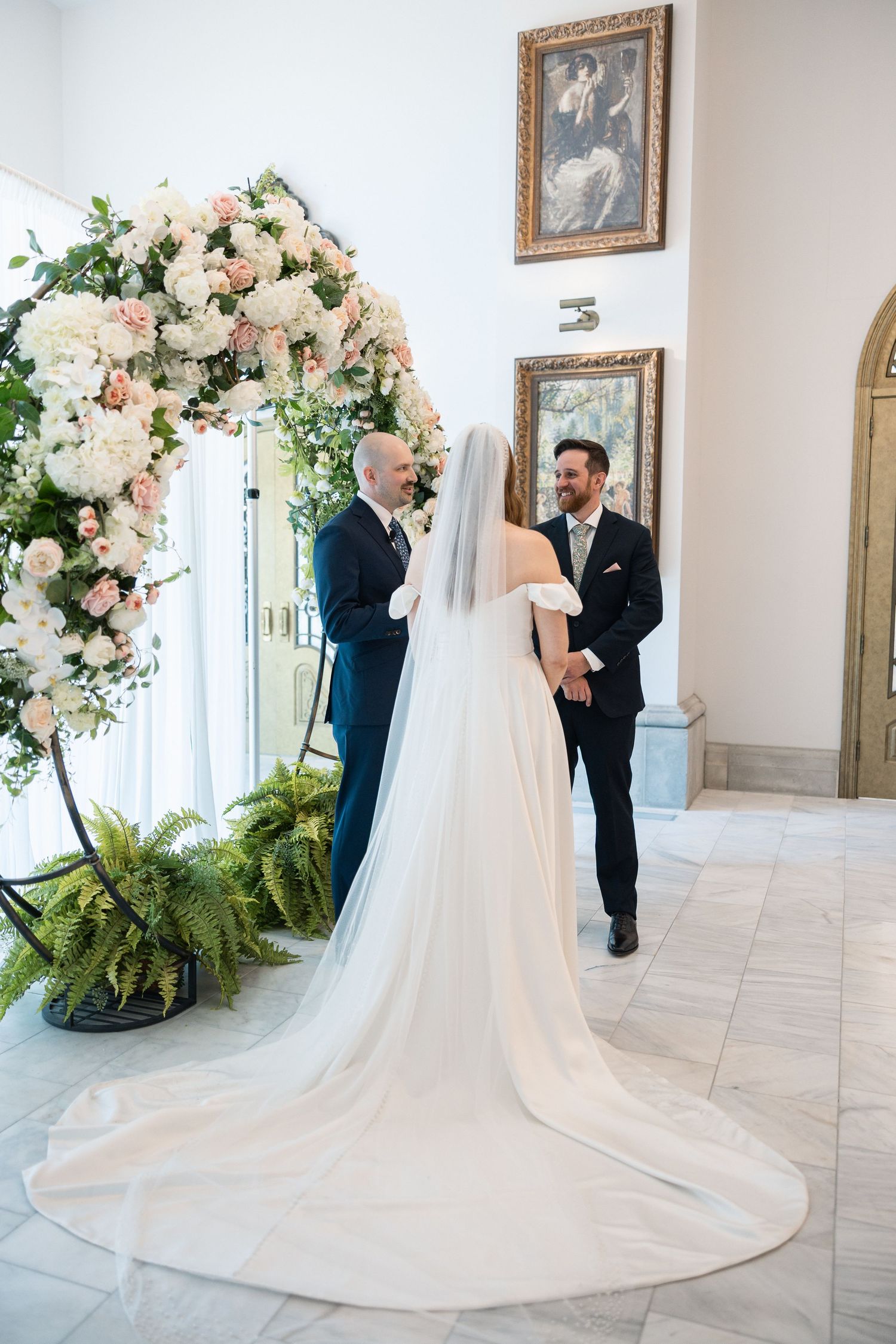Wedding ceremony setup with luxurious floral arch and flowing white dress train in an elegant white room.