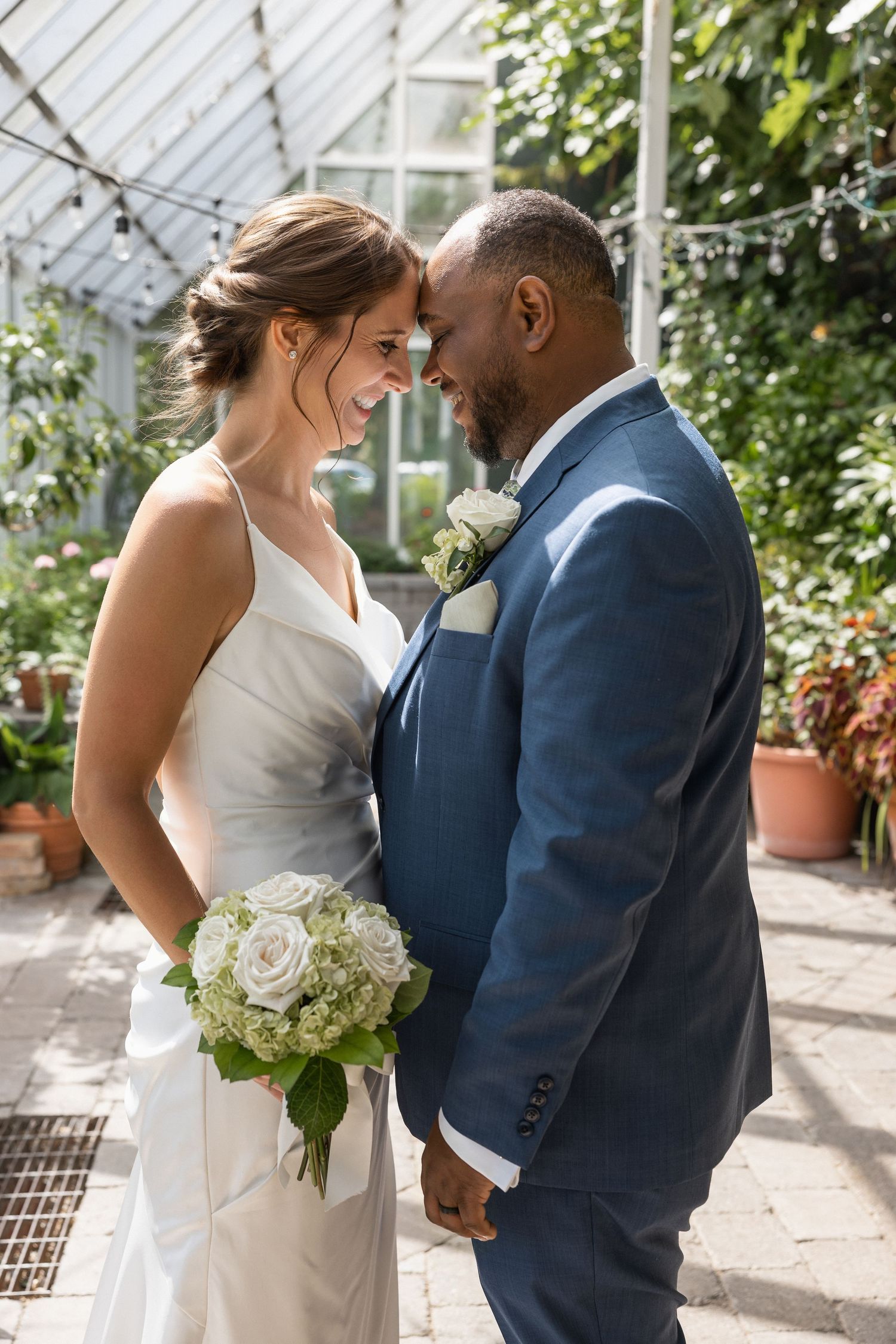 Intimate wedding portraits captured in a light-filled greenhouse with white roses and greenery.