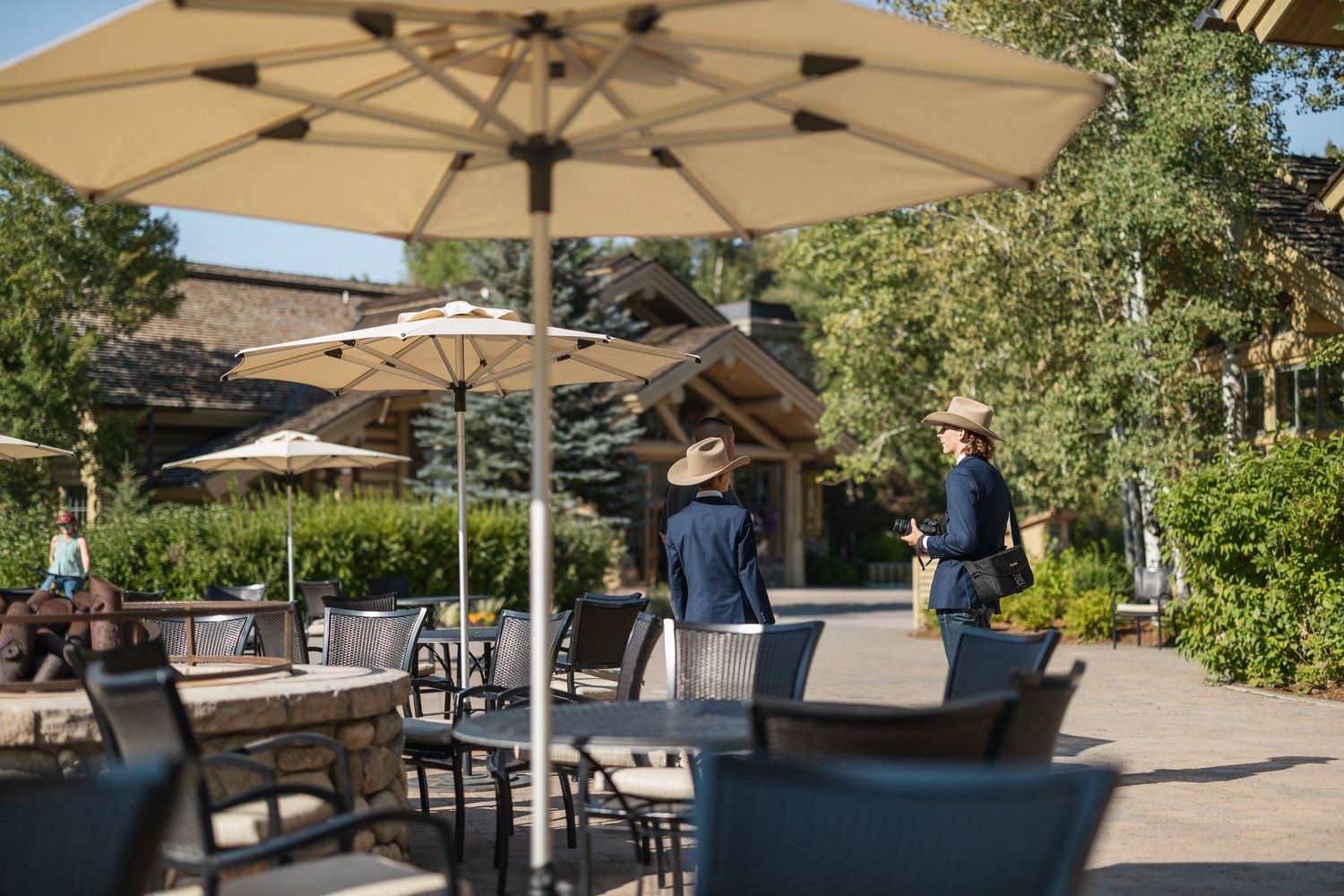 Outdoor dining area with beige umbrellas and tables set up on a stone patio surrounded by greenery.
