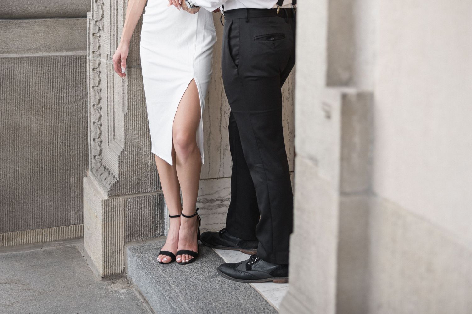 A couple in formal attire stands close together against a stone wall in an elegant architectural setting.
