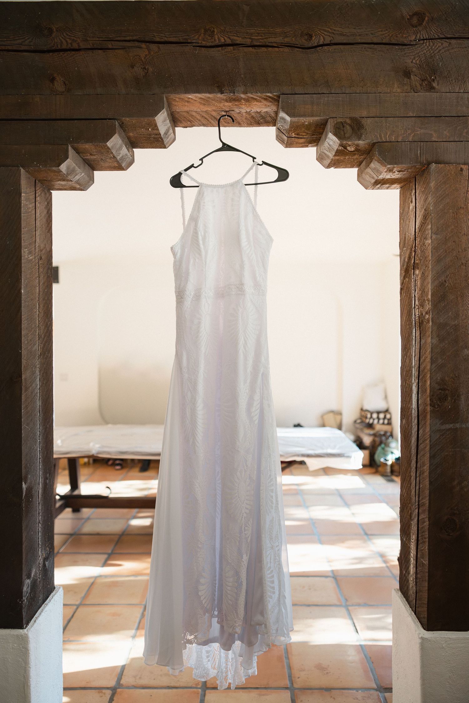 An elegant white wedding dress hangs in a rustic wooden doorframe with sunlit tile floors visible in the background.