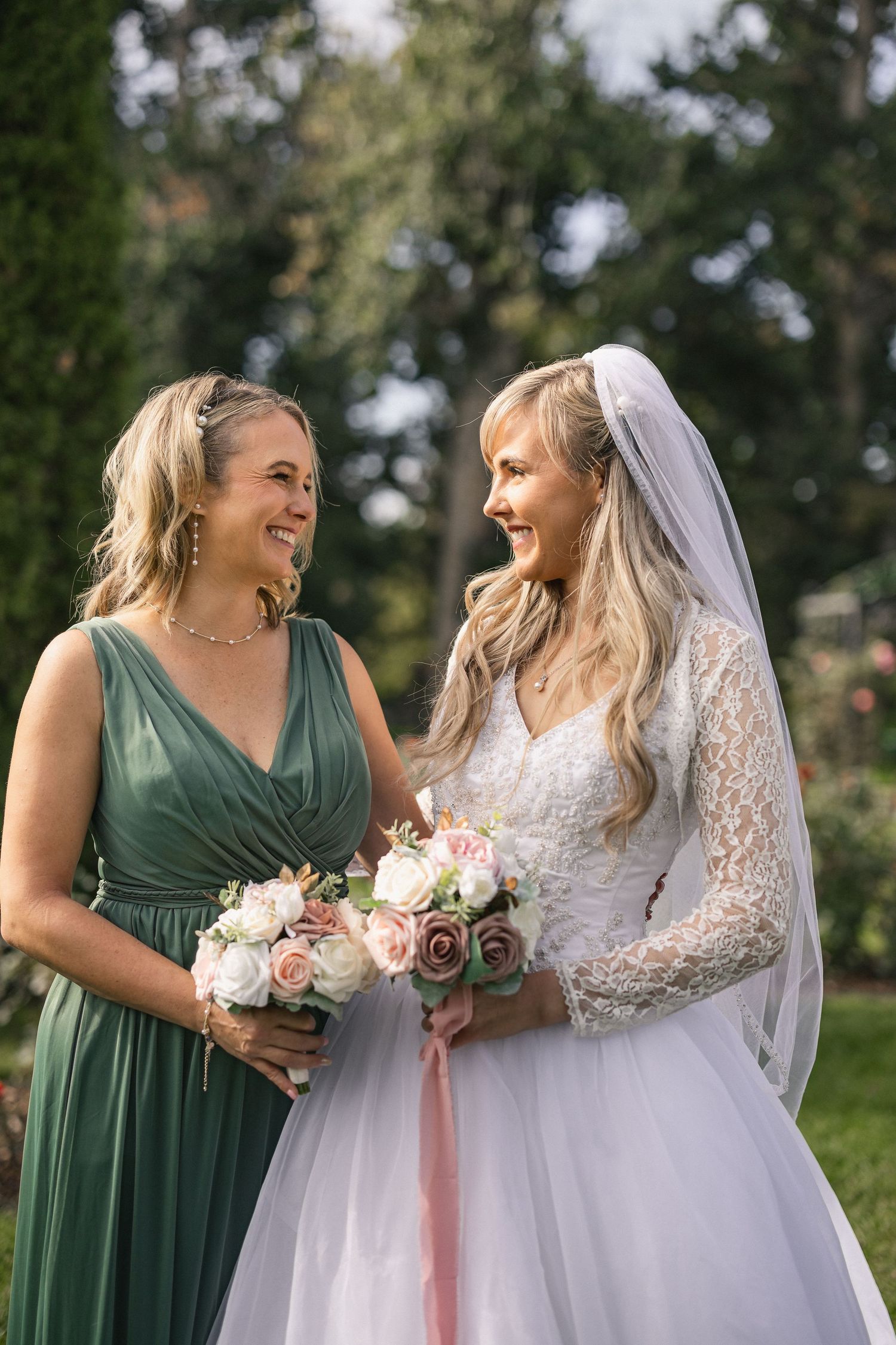 Bridesmaid in sage green dress shares a happy moment with the bride holding matching floral bouquets.