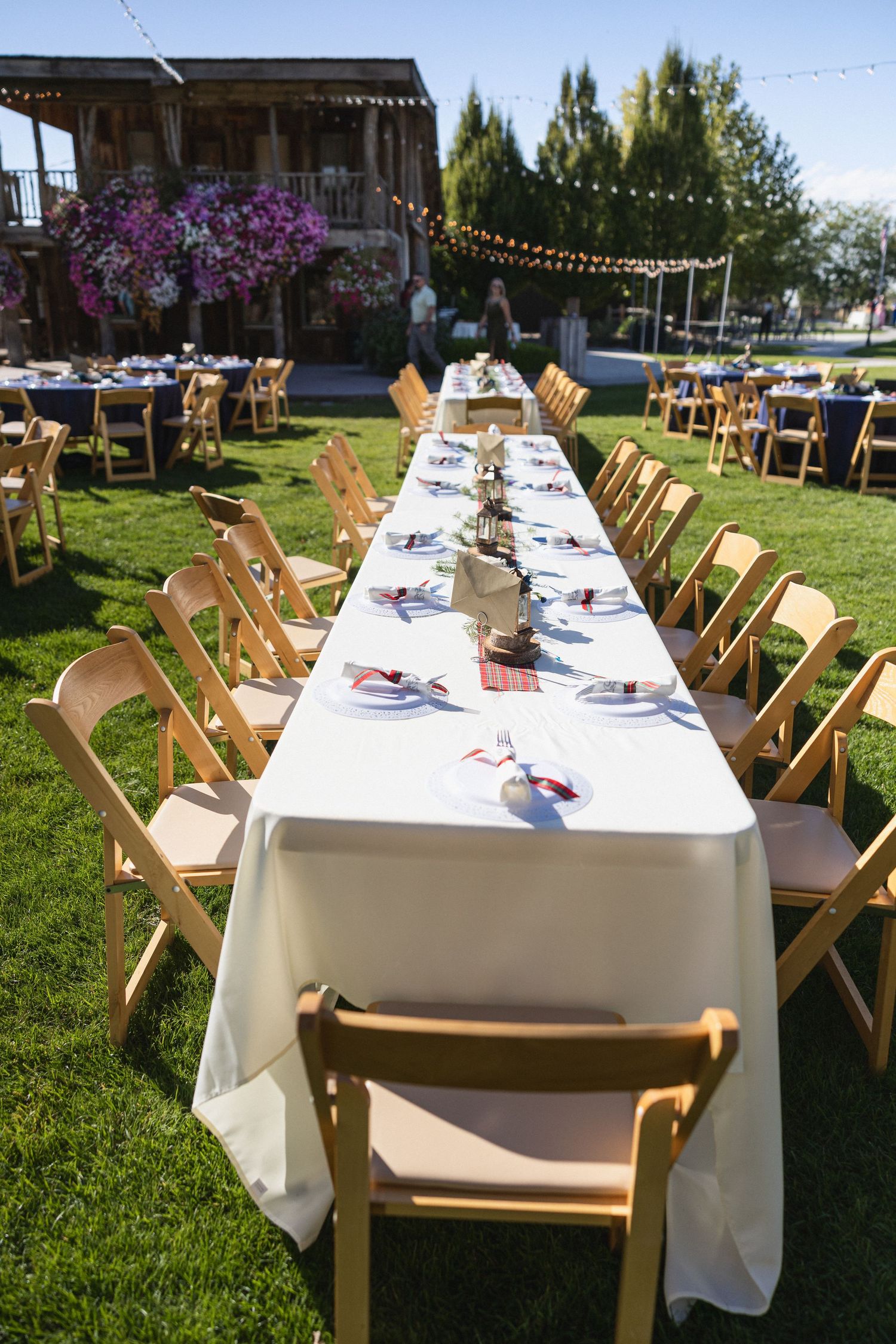 Outdoor wedding reception setup with long white tables and wooden chairs under string lights.