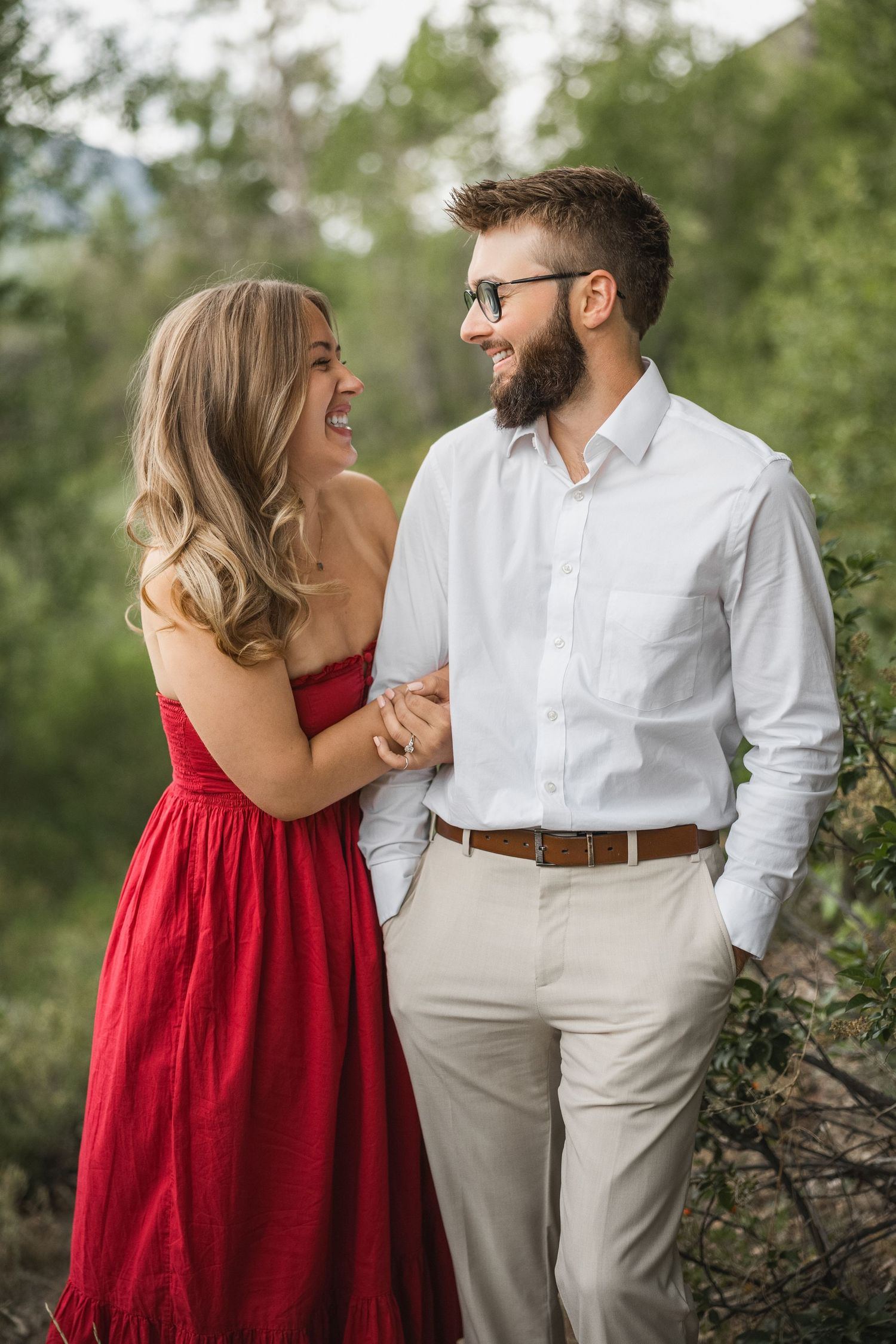 A couple shares an intimate moment outdoors, with the woman in a red dress and man in white shirt and khaki pants.