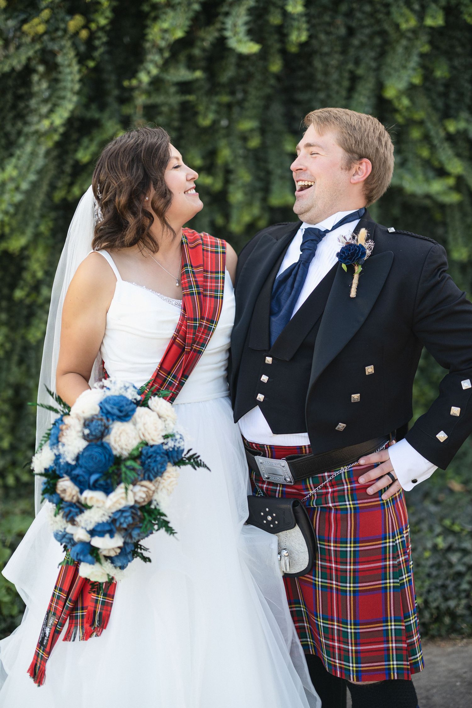 Scottish-themed wedding couple share a laugh against ivy wall backdrop.