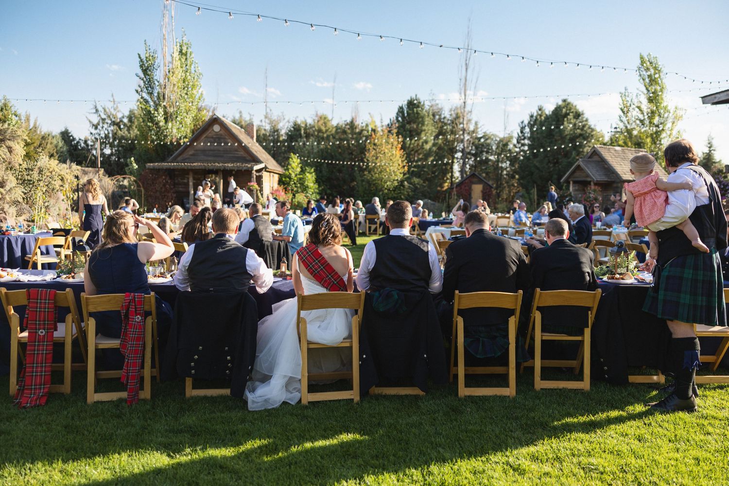 Wedding guests dining at long tables during outdoor reception with church in background.