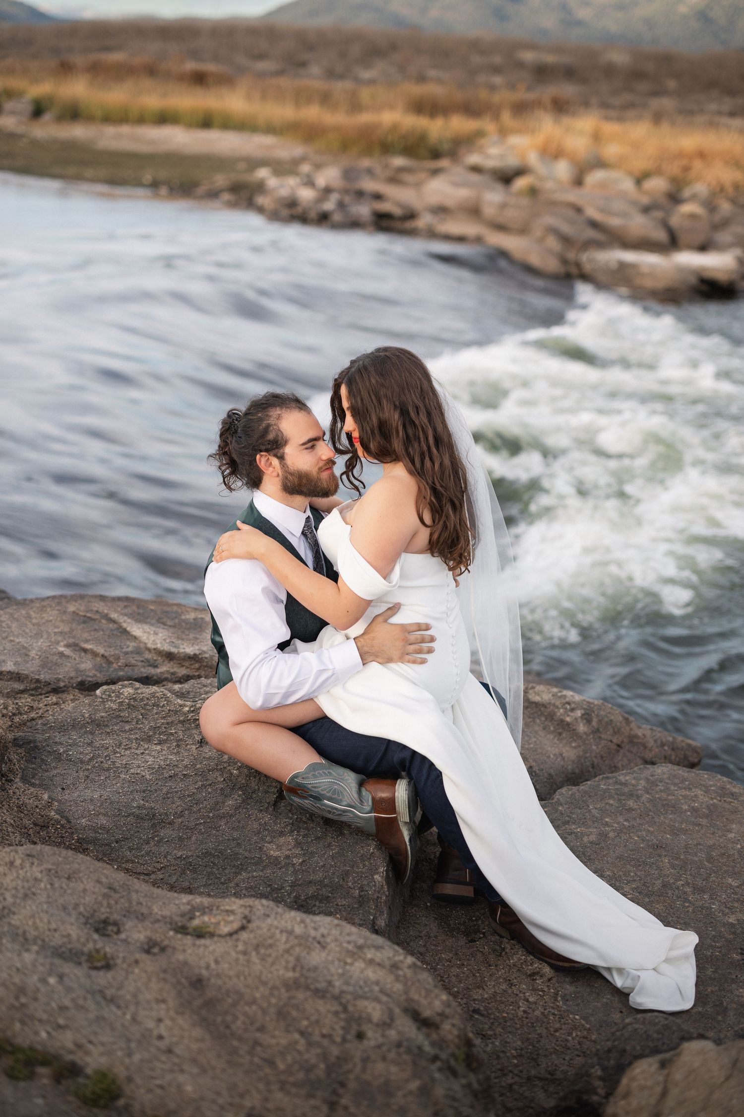 Wedding photoshoot on rocky riverbank with flowing water and mountain scenery.