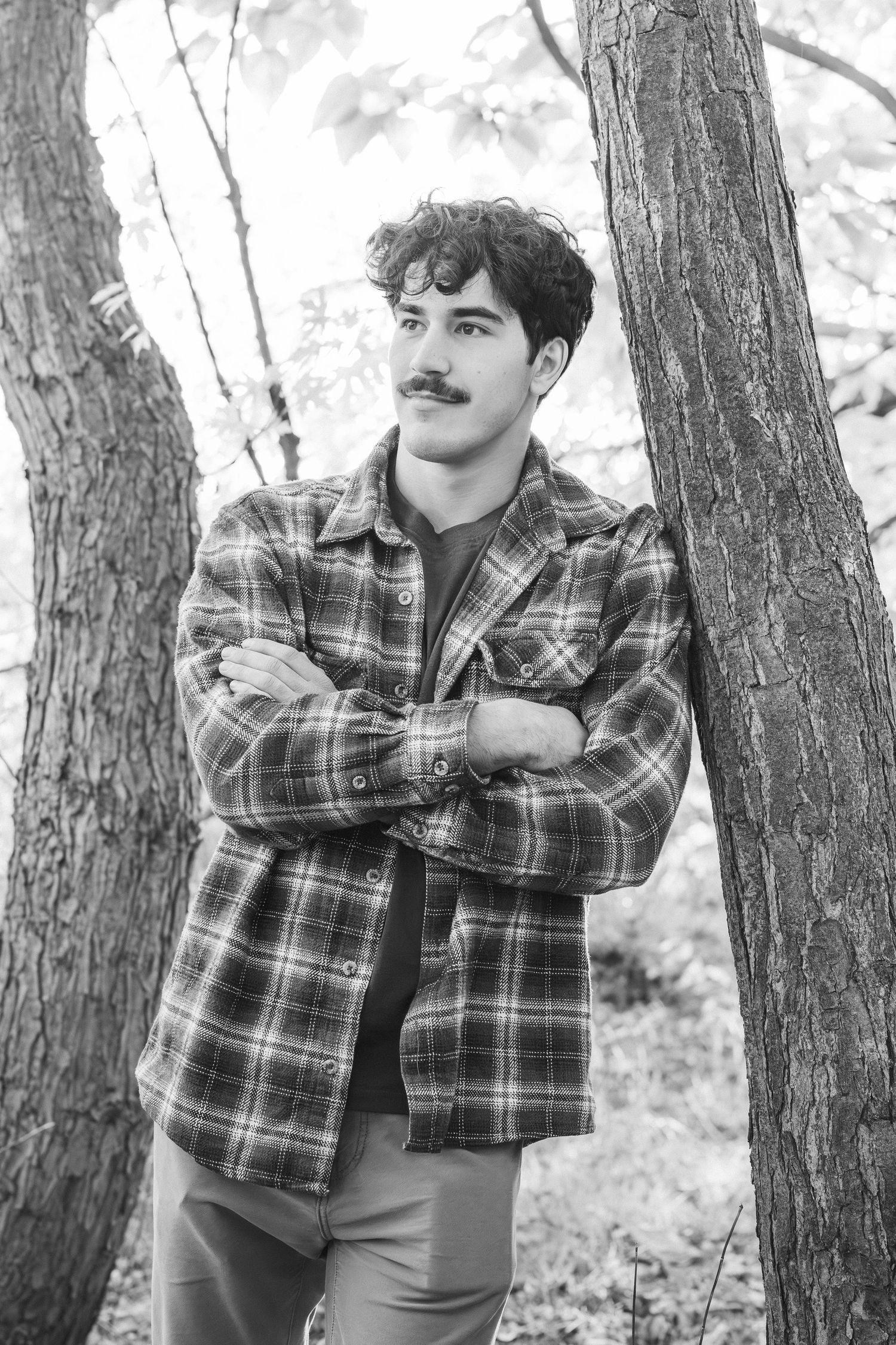 Black and white photo of person in plaid shirt posing against tree trunks.