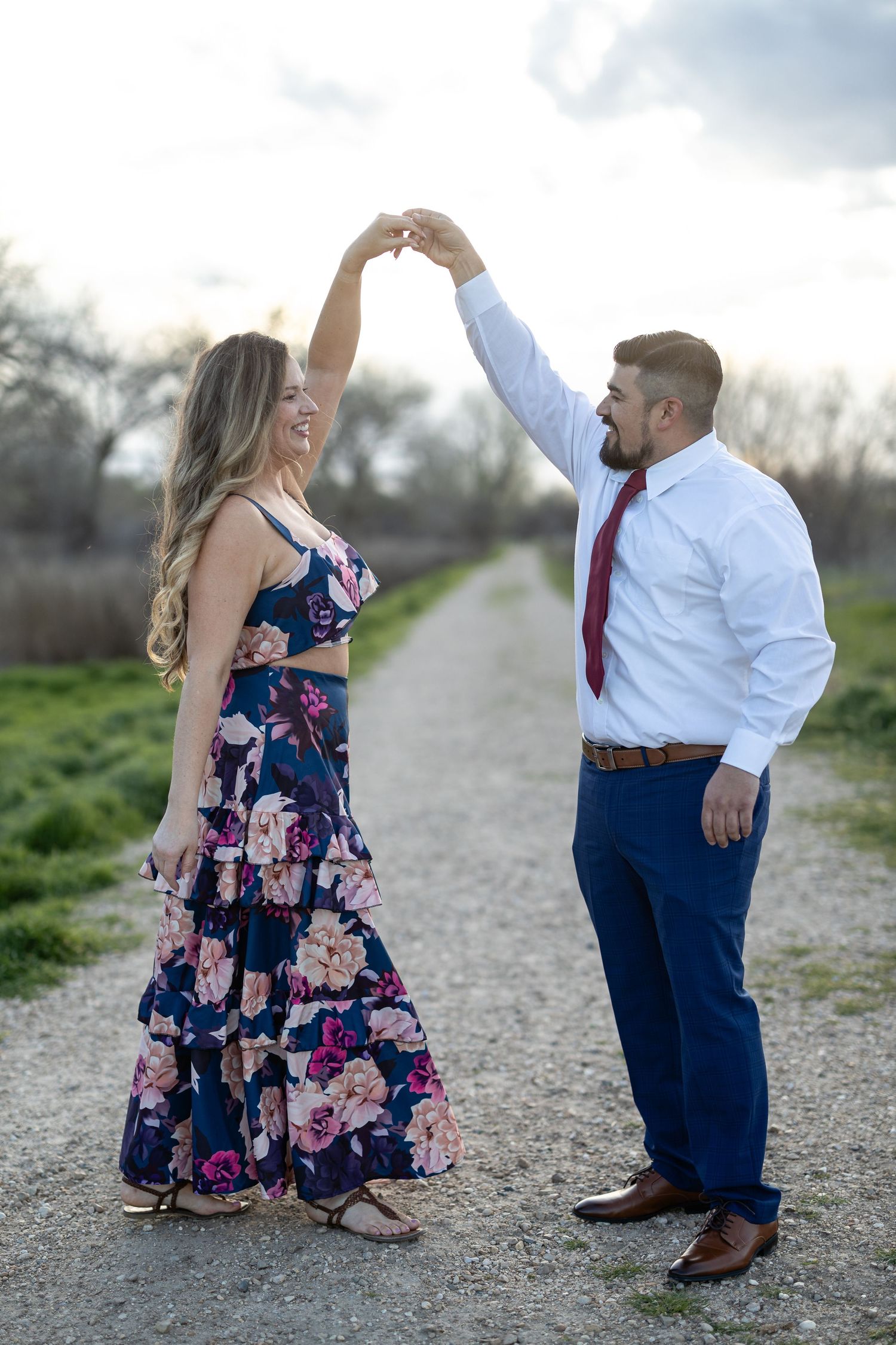 A couple dancing together on a path with the woman wearing a floral maxi dress and the man in business attire.