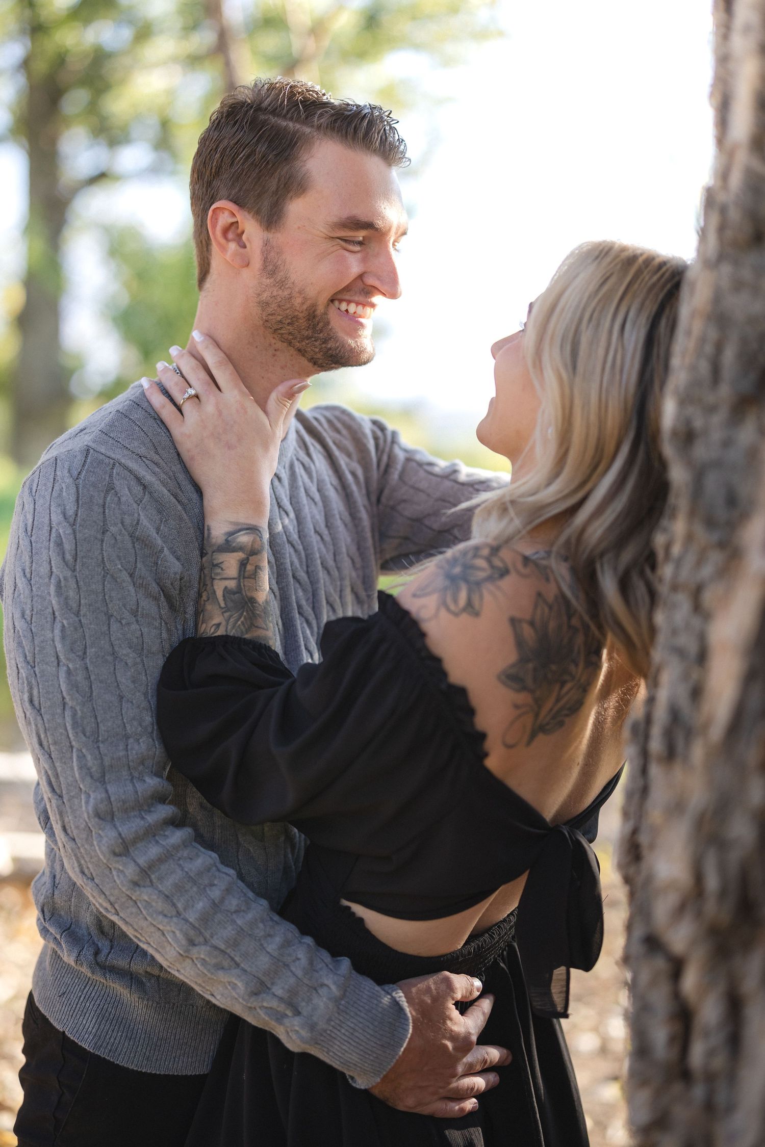 Couple shares tender moment outdoors wearing gray sweater and black dress with visible tattoos.