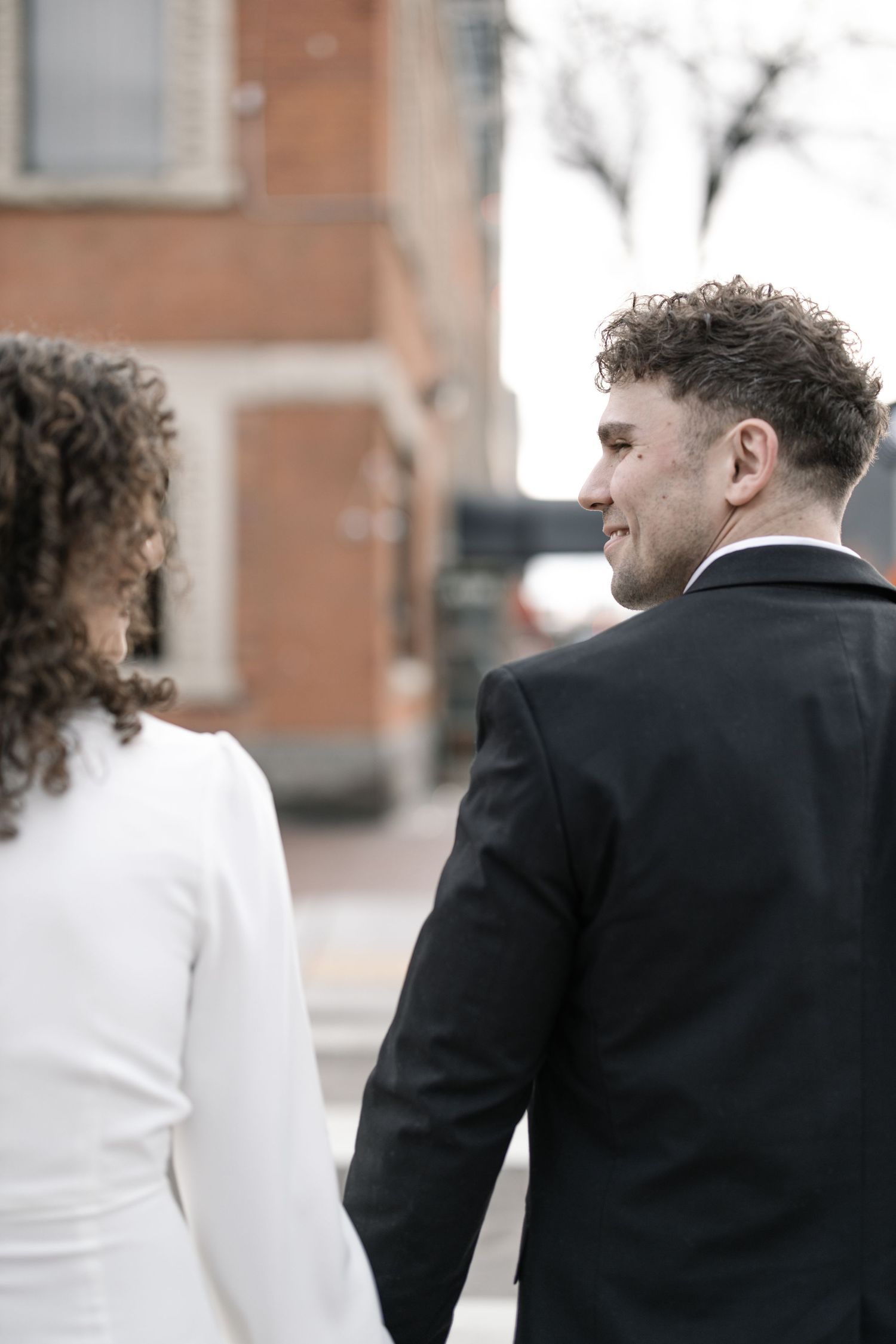 A couple in formal wear walks together near brick buildings in an urban setting.