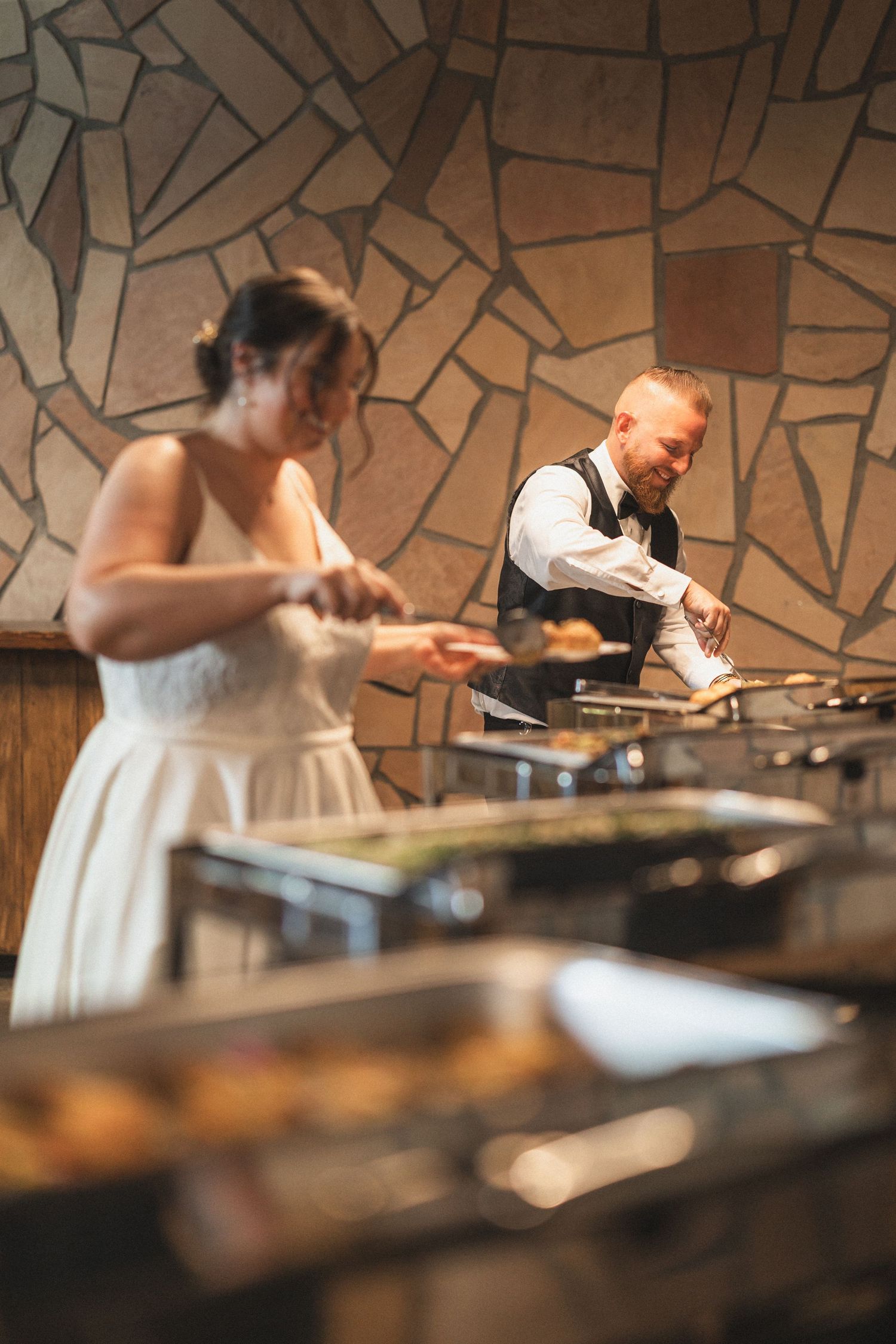 Newlyweds serving food at their wedding buffet with stone wall backdrop.