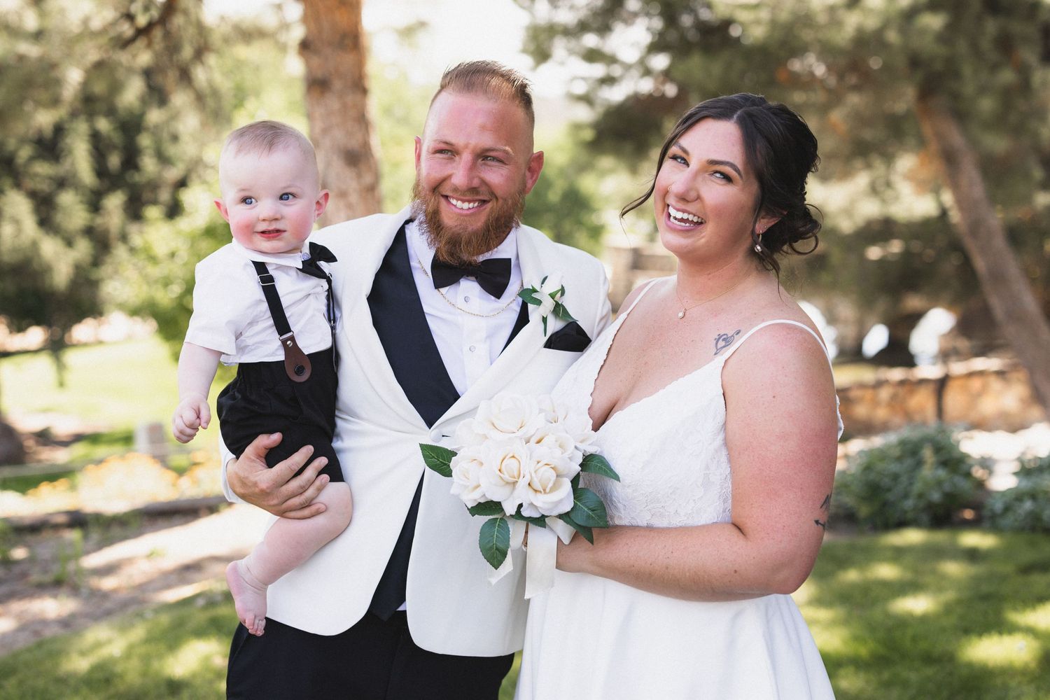 A joyful wedding portrait in a garden setting featuring a couple in white attire with a small child in formal wear.