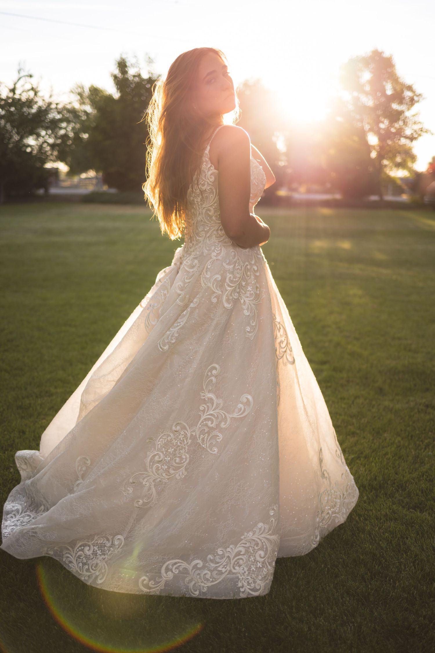 A bride in a lace wedding gown poses in a sunlit field during golden hour.