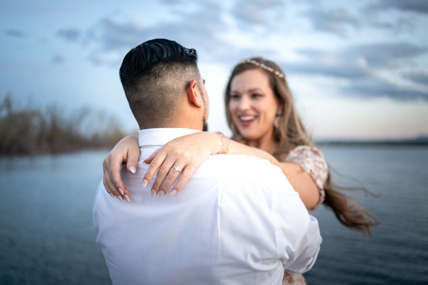 A romantic embrace captured on a lakeside dock during sunset with dramatic clouds.