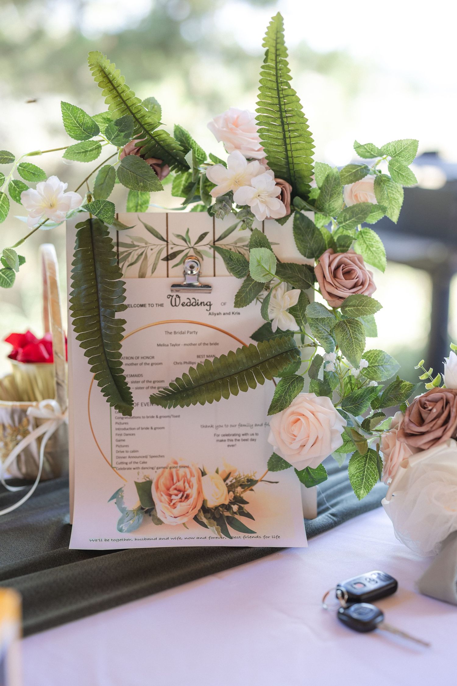 A wedding program decorated with ferns and roses sits among floral arrangements.