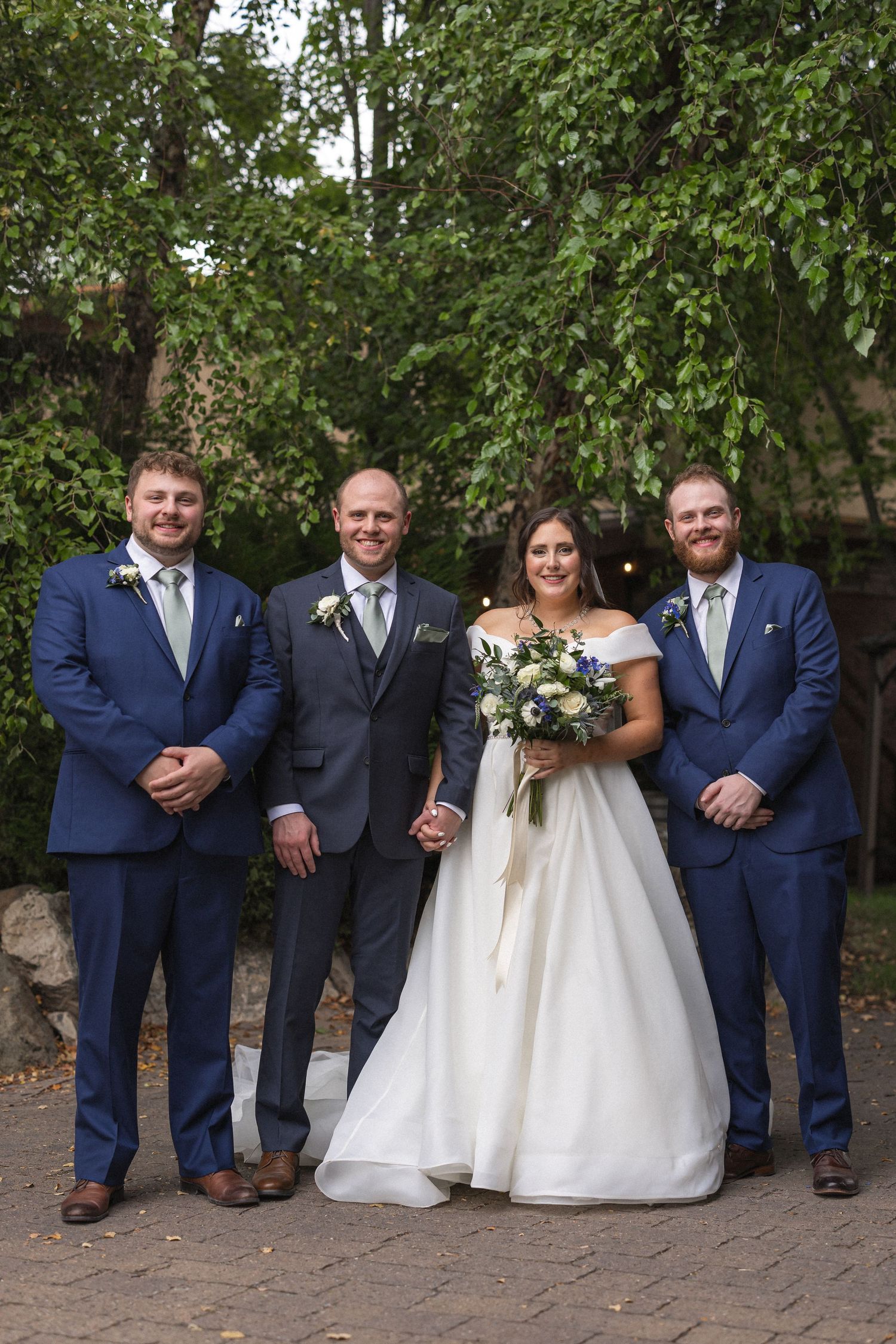 Wedding party members in navy and grey suits pose with couple on stone pathway.