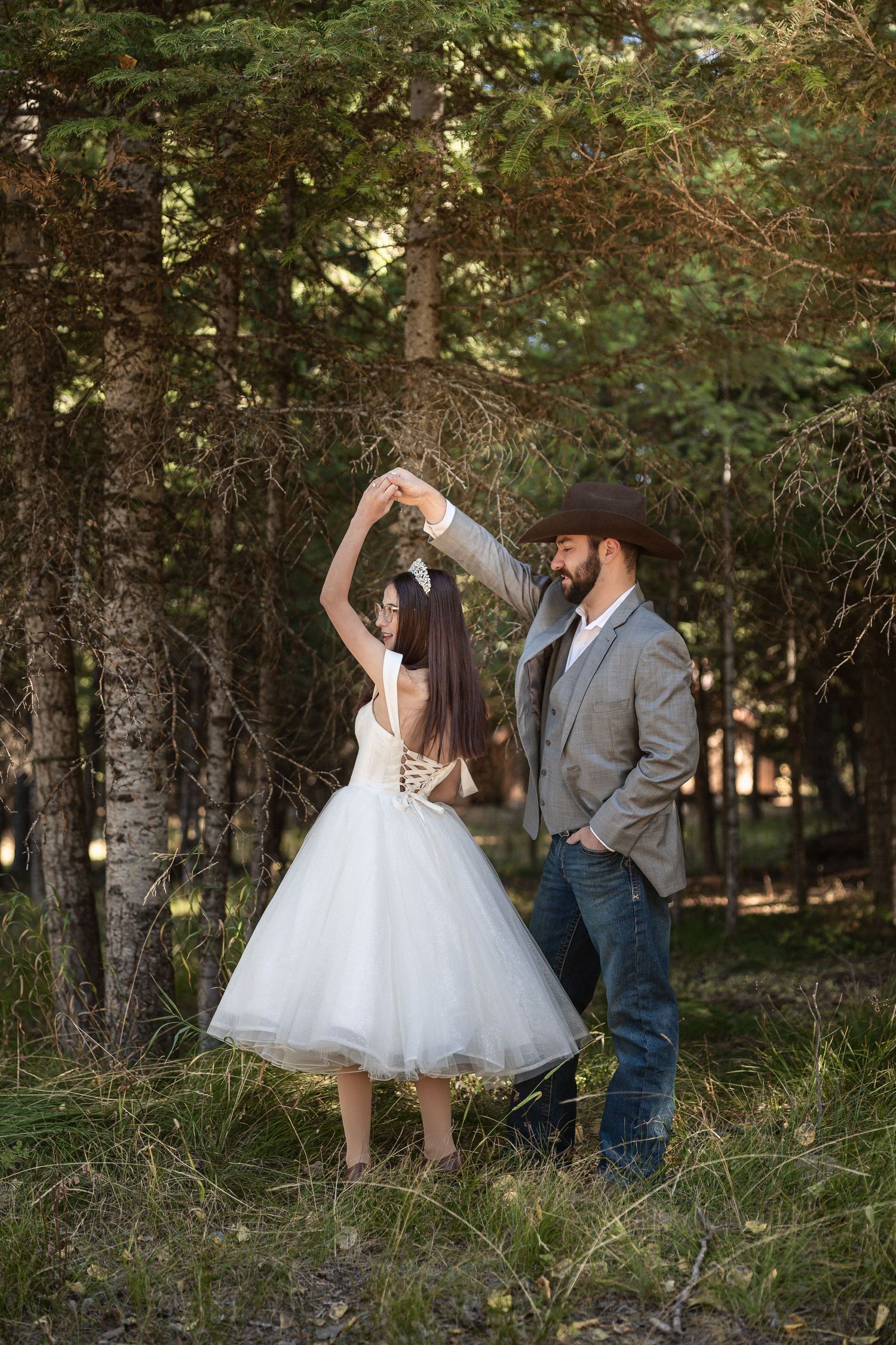 A couple dancing in a forest with bride in tea-length dress and groom in cowboy hat.