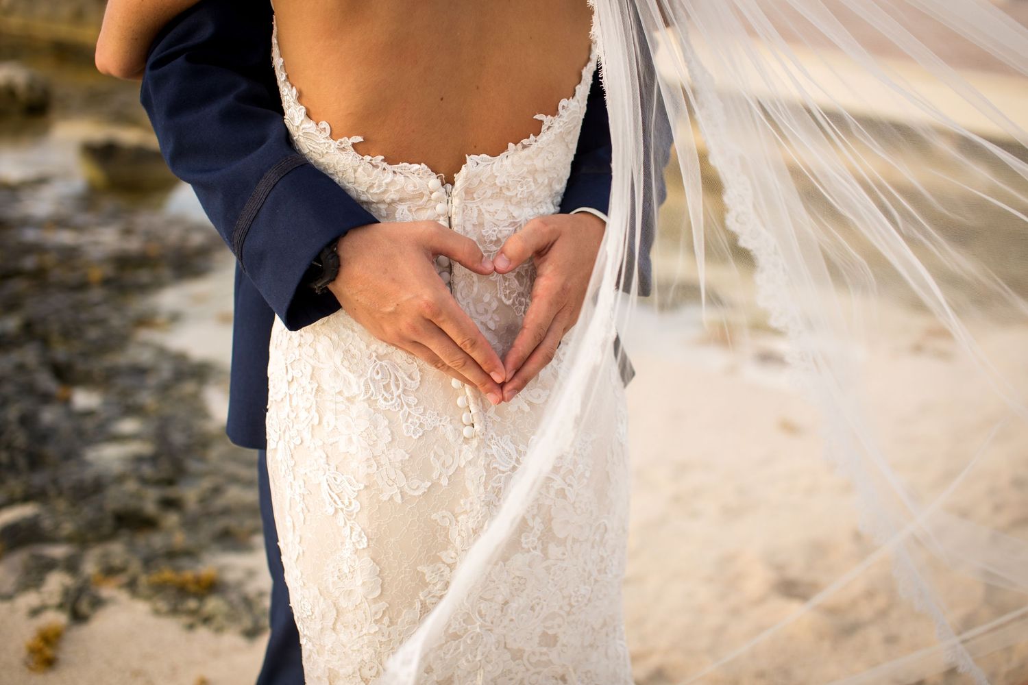 A close-up of hands forming a heart shape against a white lace wedding dress and navy suit.