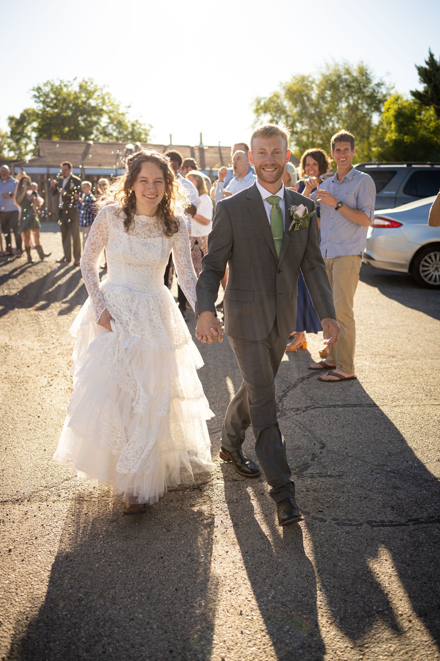 Wedding couple walks together hand in hand on a sunny day while guests look on in the background.