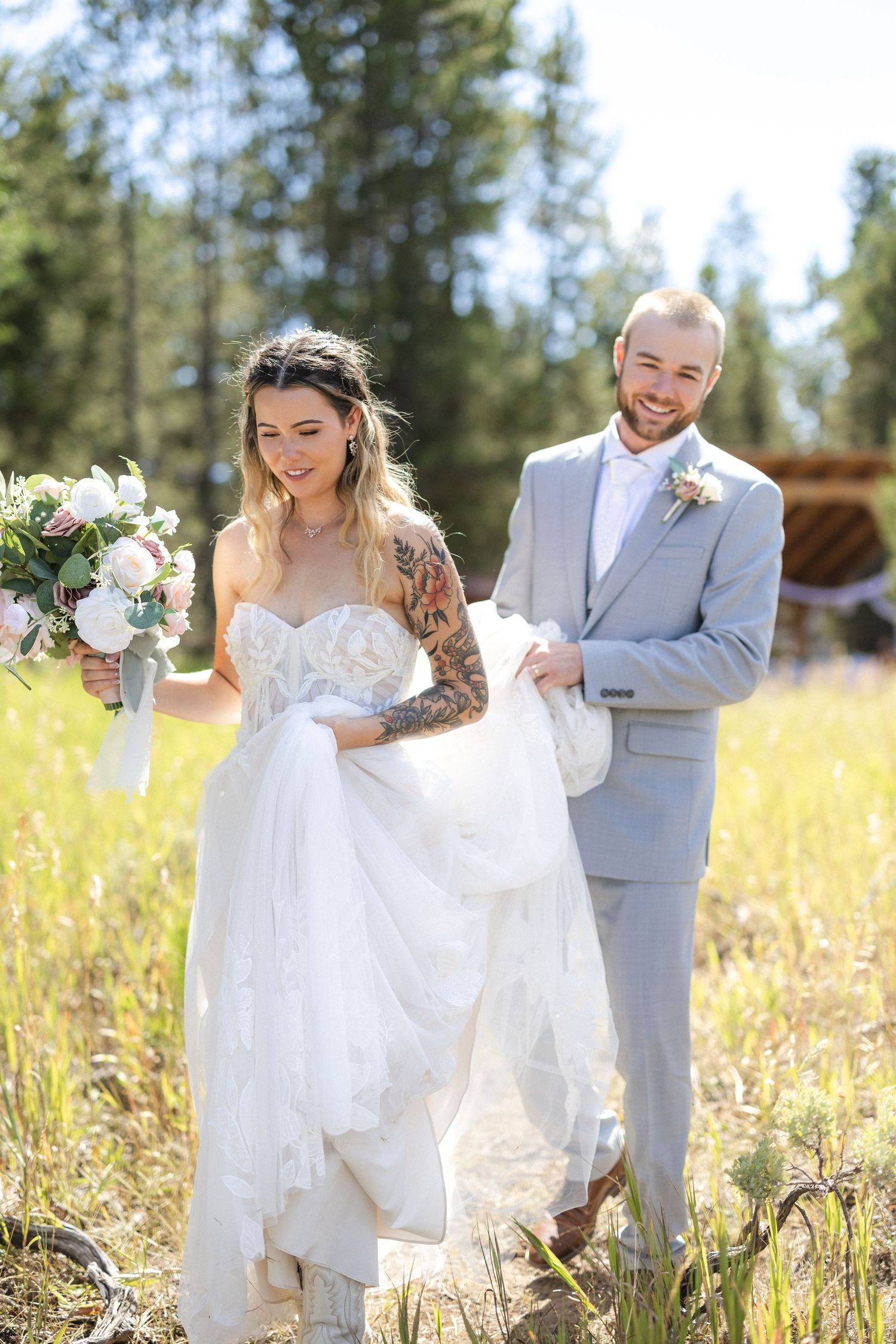 A couple poses playfully in a golden field during a wedding photoshoot near pine trees.