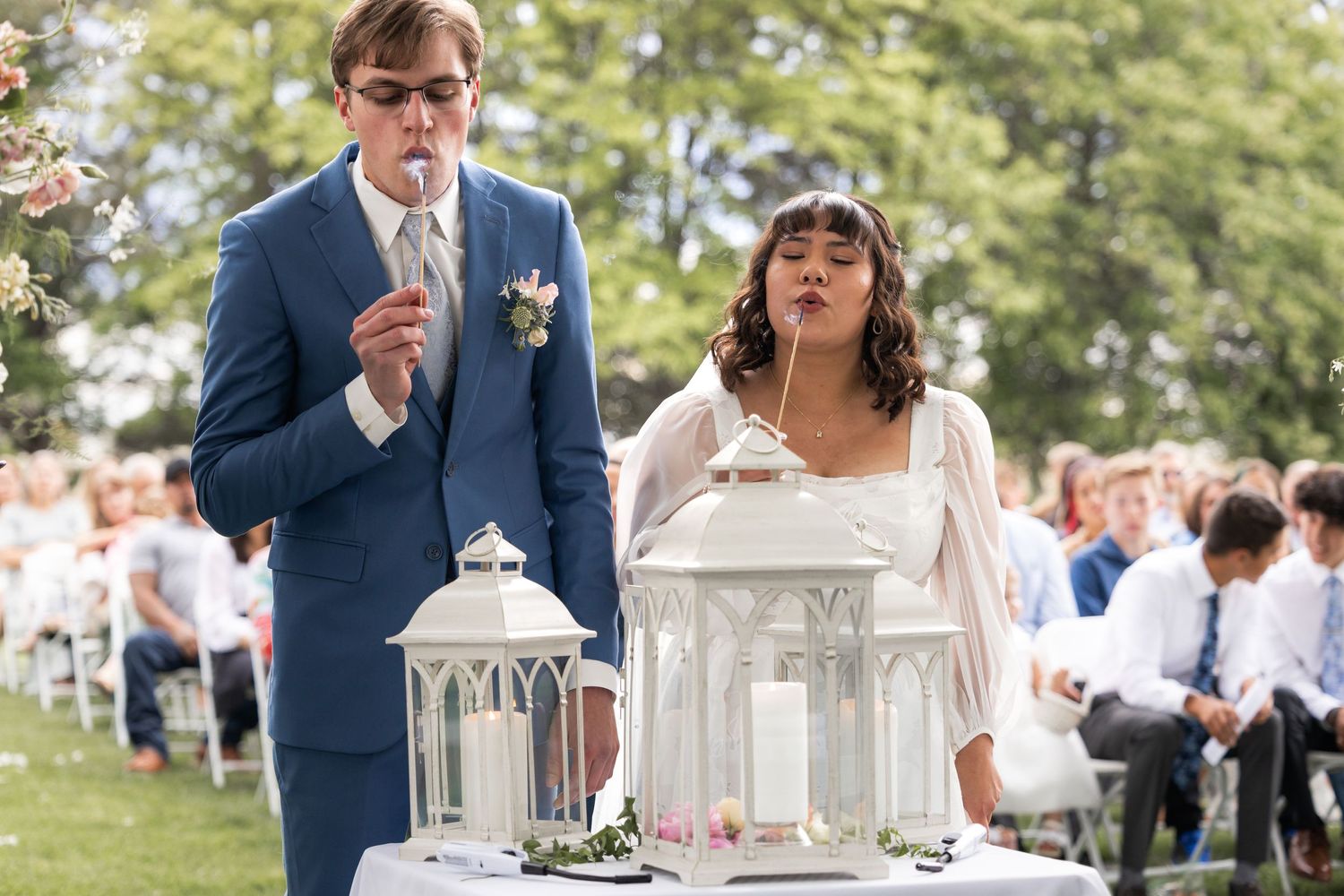 Wedding couple examining decorative lanterns on white-clothed table during outdoor ceremony.