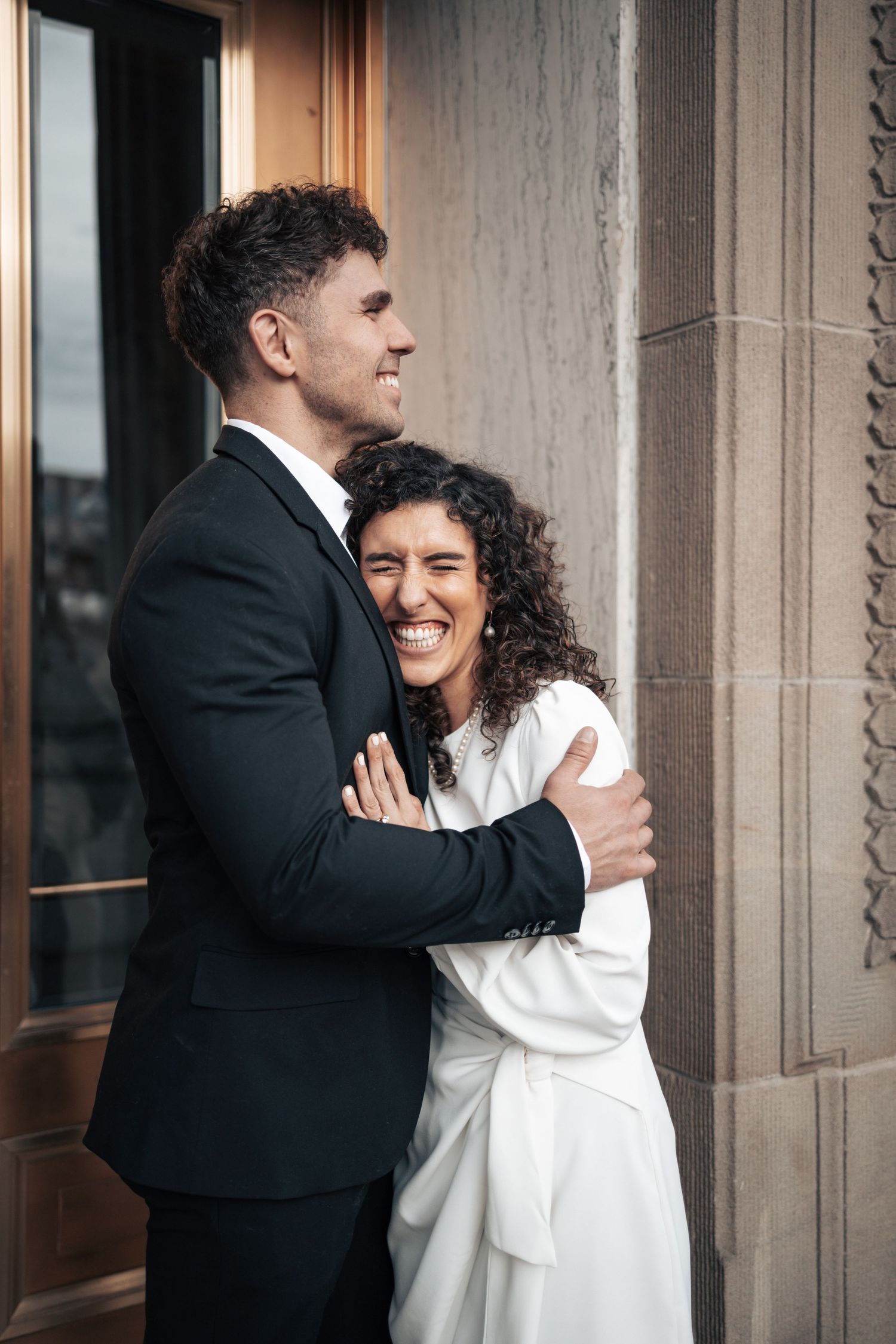 A couple in black and white attire share a warm embrace near architectural columns.