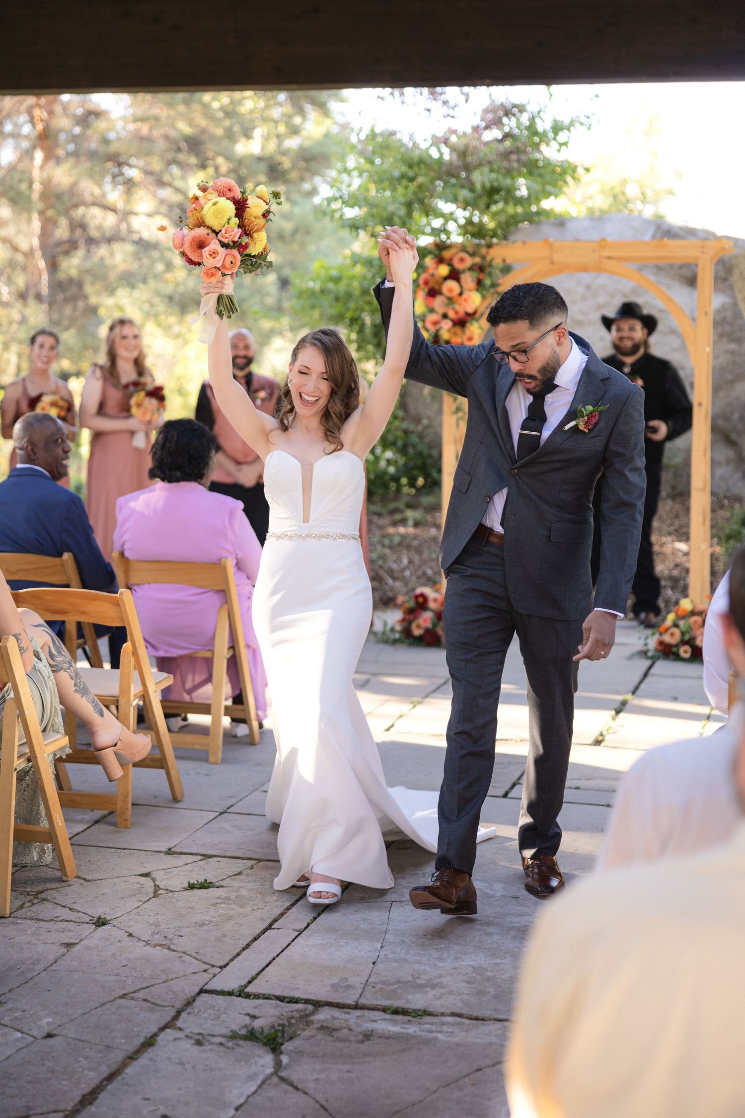 Newlyweds celebrate with raised arms while walking down the aisle at an outdoor wedding ceremony.