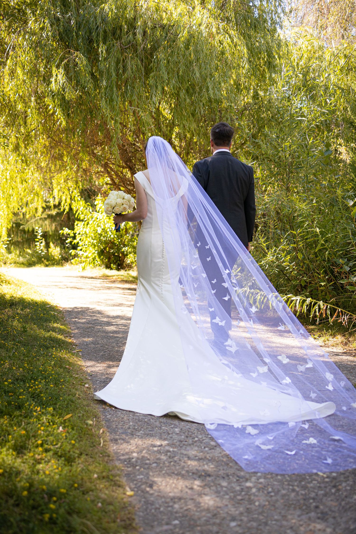 Bride's long cathedral veil with butterfly details trails behind couple on garden path.