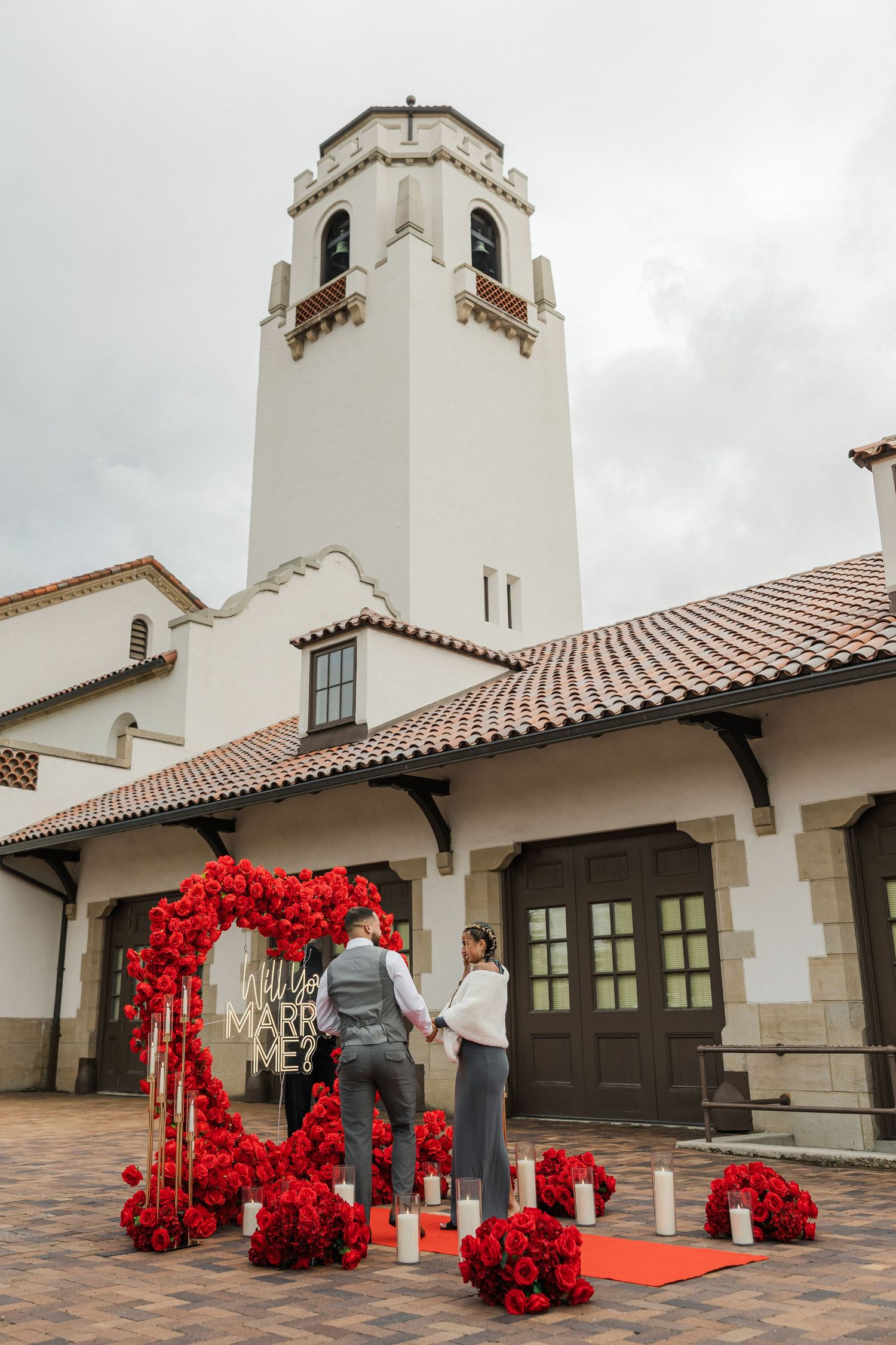 Couple's romantic marriage proposal with red roses heart arch and 'Marry Me' sign outside a white Spanish-style church.