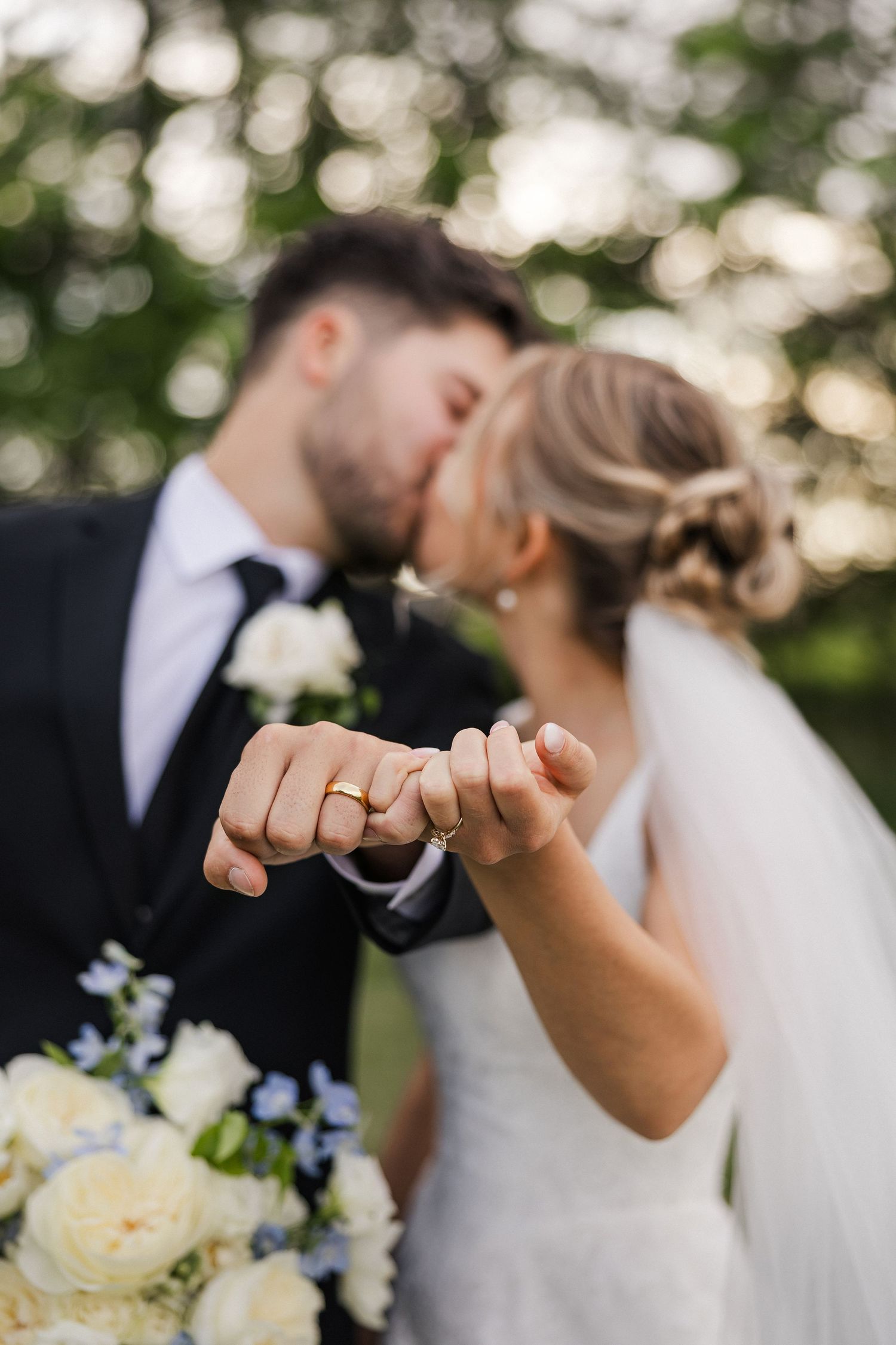 Newlyweds kissing while showing off their wedding rings, with a white floral bouquet in an outdoor setting.