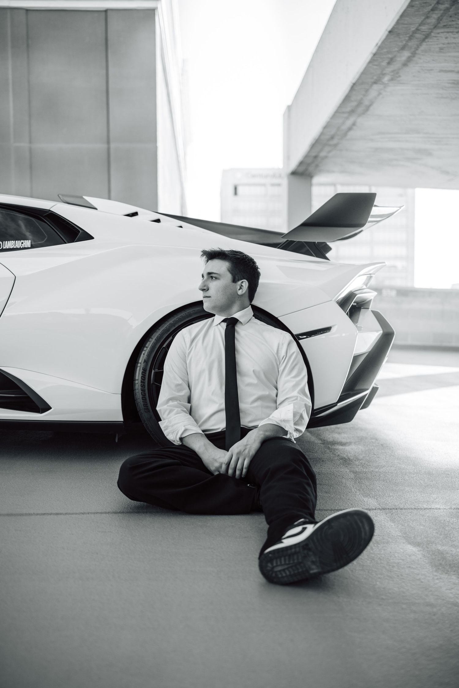 Black and white photo of someone in formal wear sitting beside sleek sports car under concrete structure.
