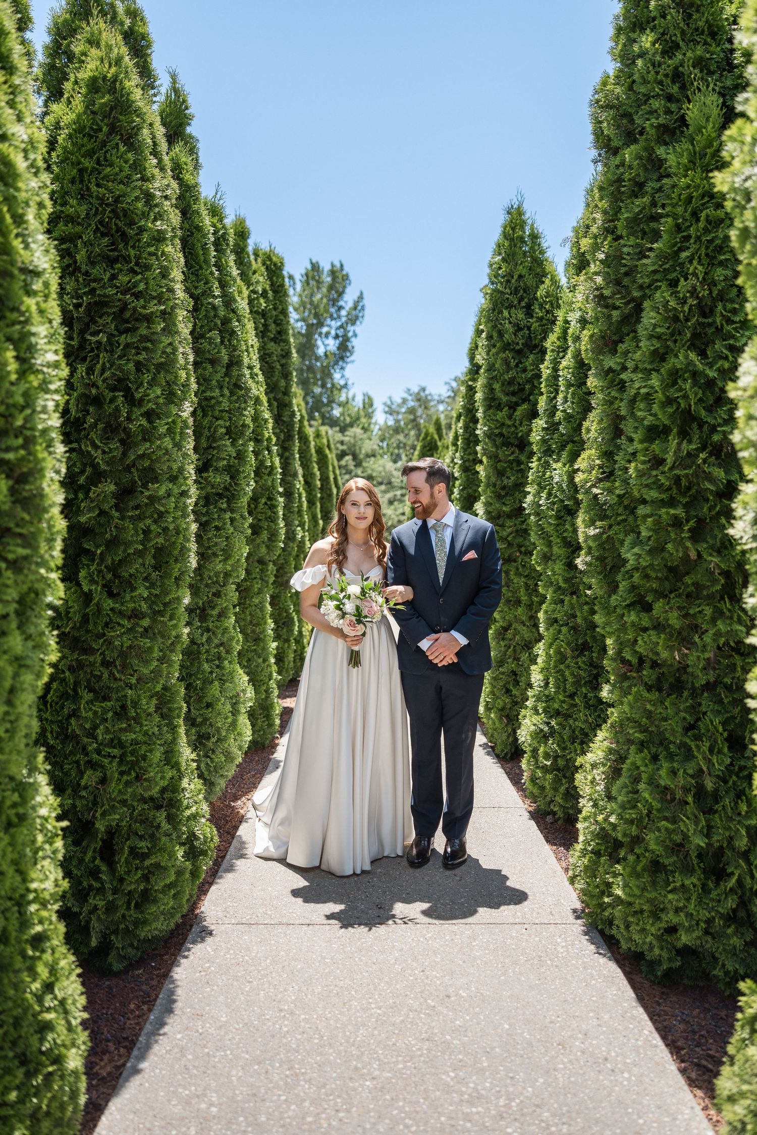 Wedding couple standing in beautiful garden path lined with tall cypress trees.