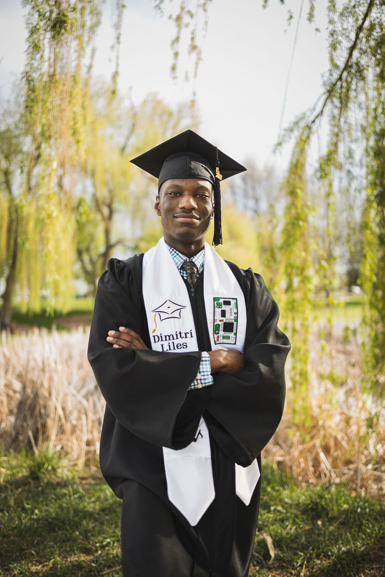 Graduate in black cap and gown with white stole poses confidently against backdrop of willow trees and tall grass.