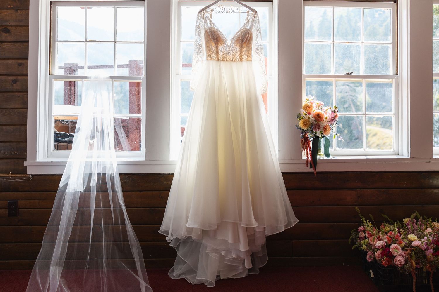 A white wedding dress and veil hang elegantly in front of sunlit windows with a floral bouquet nearby.