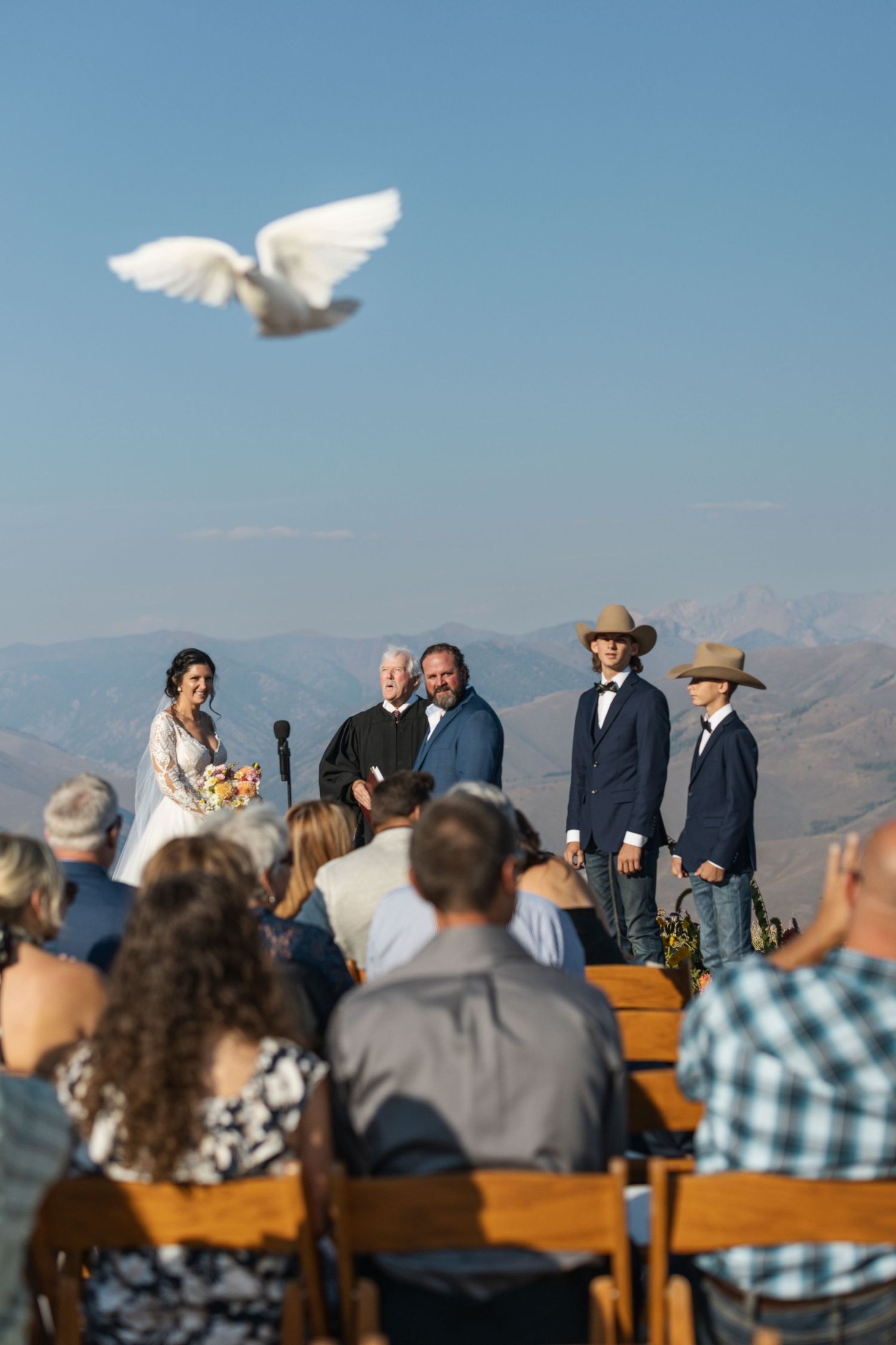 Outdoor wedding ceremony with wooden benches and mountain vista backdrop on a clear sunny day.