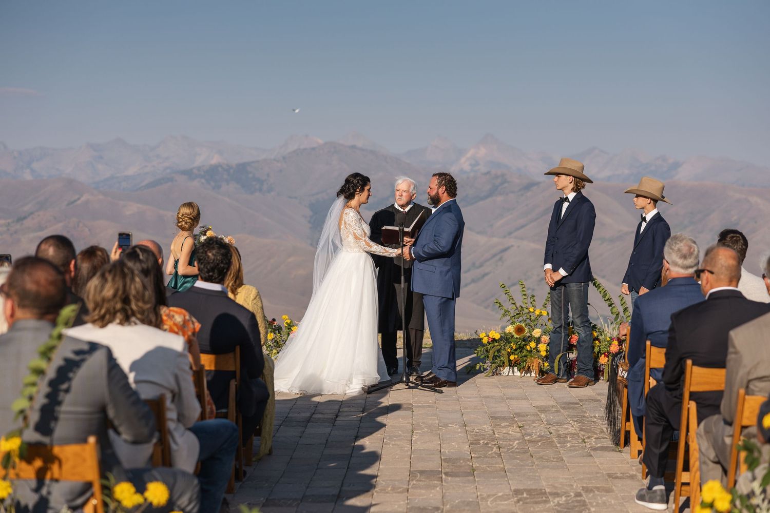 A scenic mountain wedding ceremony takes place on a stone patio with yellow flowers and mountain peaks in the background.